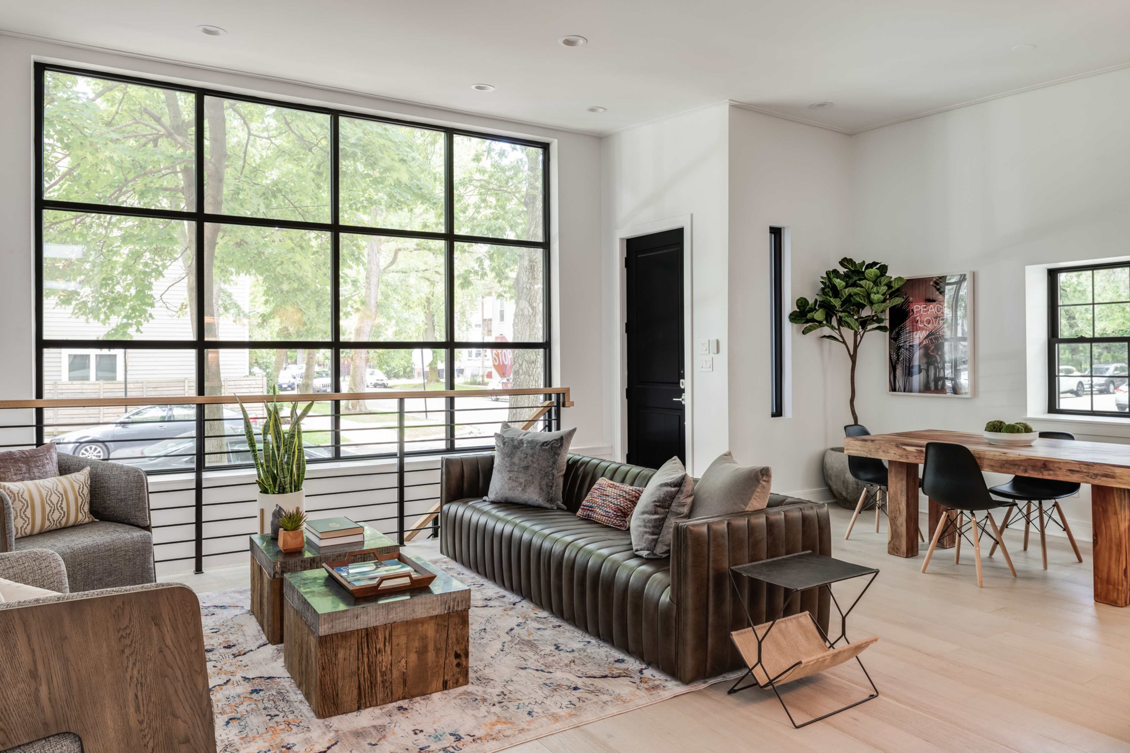 A modern living room features a large window, a leather sofa, wooden tables, and a dining area with a mix of seating styles.