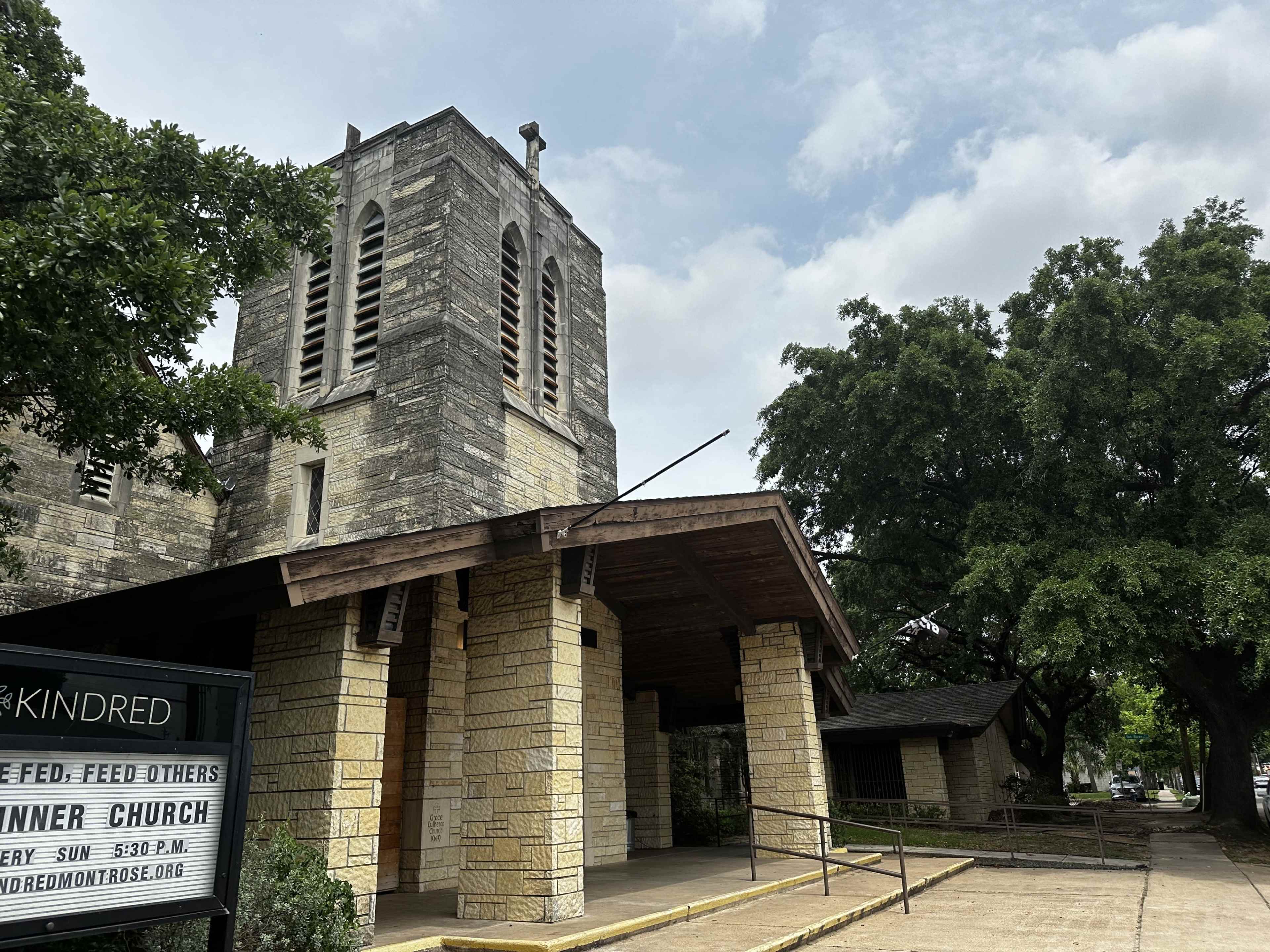 A stone church building with a tall bell tower and large trees surrounding it.