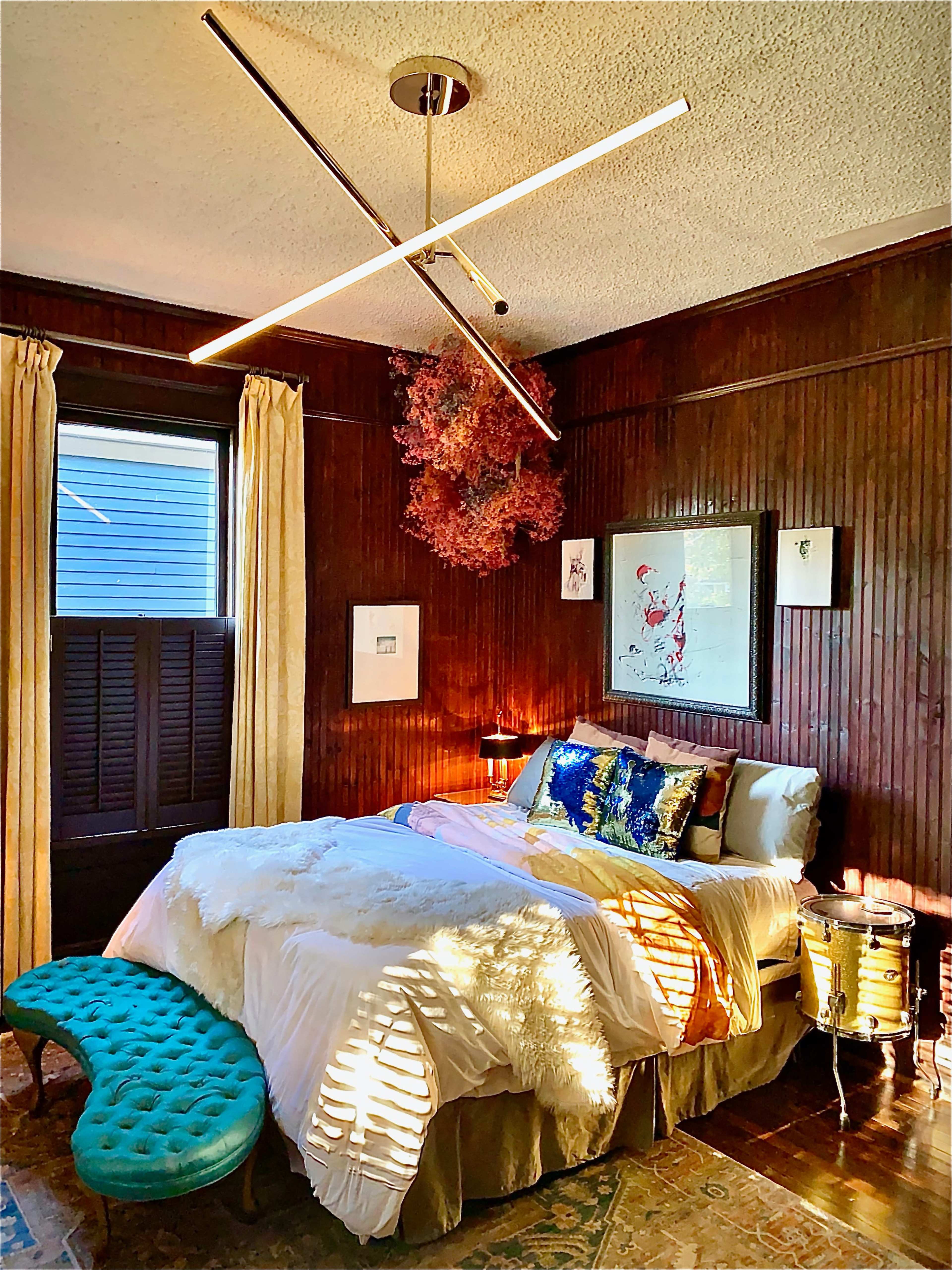 A cozy bedroom with a bed adorned with colorful pillows, a unique ceiling light fixture, and wooden paneling on the walls, complemented by curtains and a small upholstered bench at the foot of the bed.