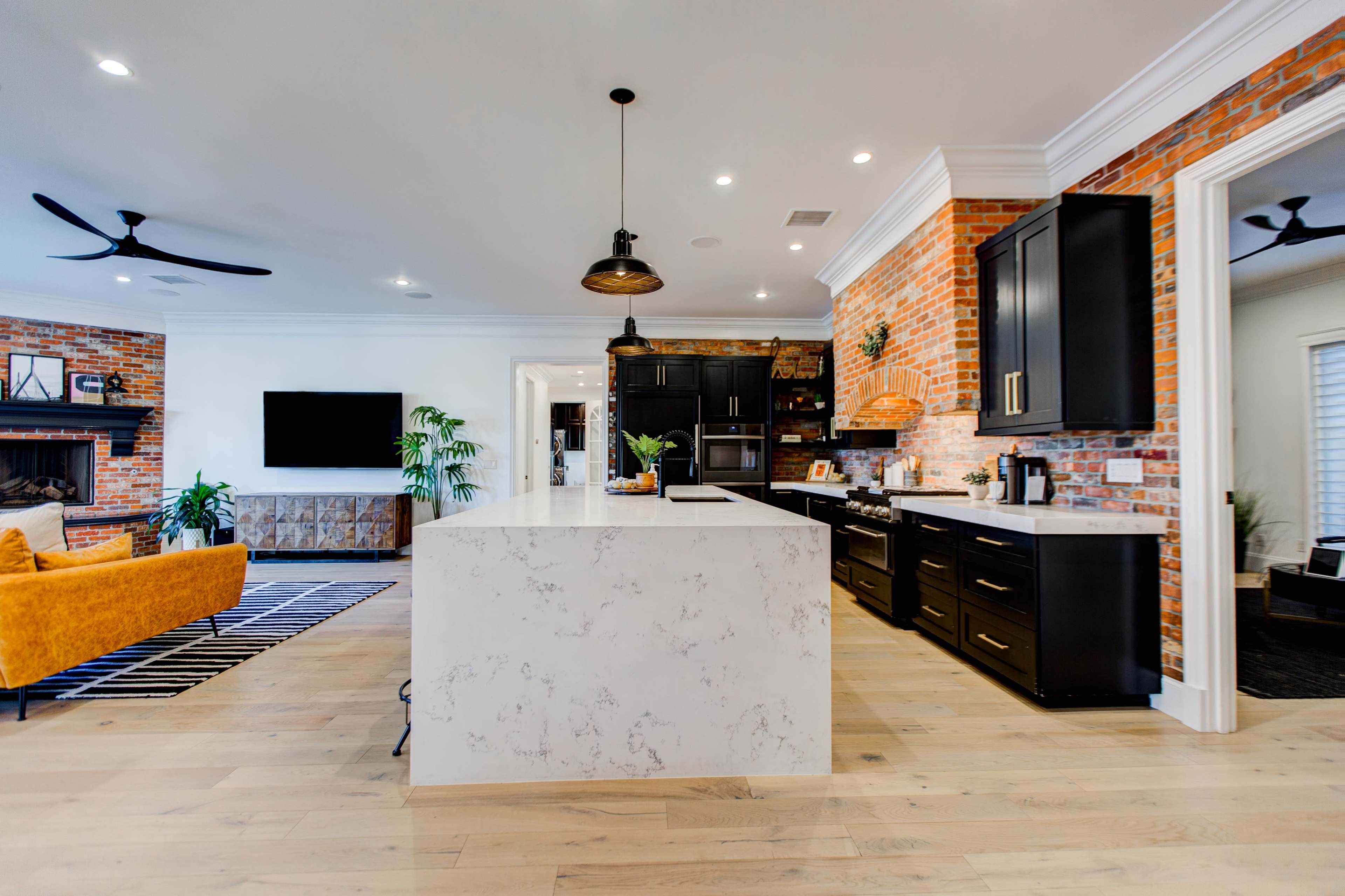 The kitchen features a large marble island with black cabinetry and a brick accent wall, overlooking a living area with a television and a yellow sofa.