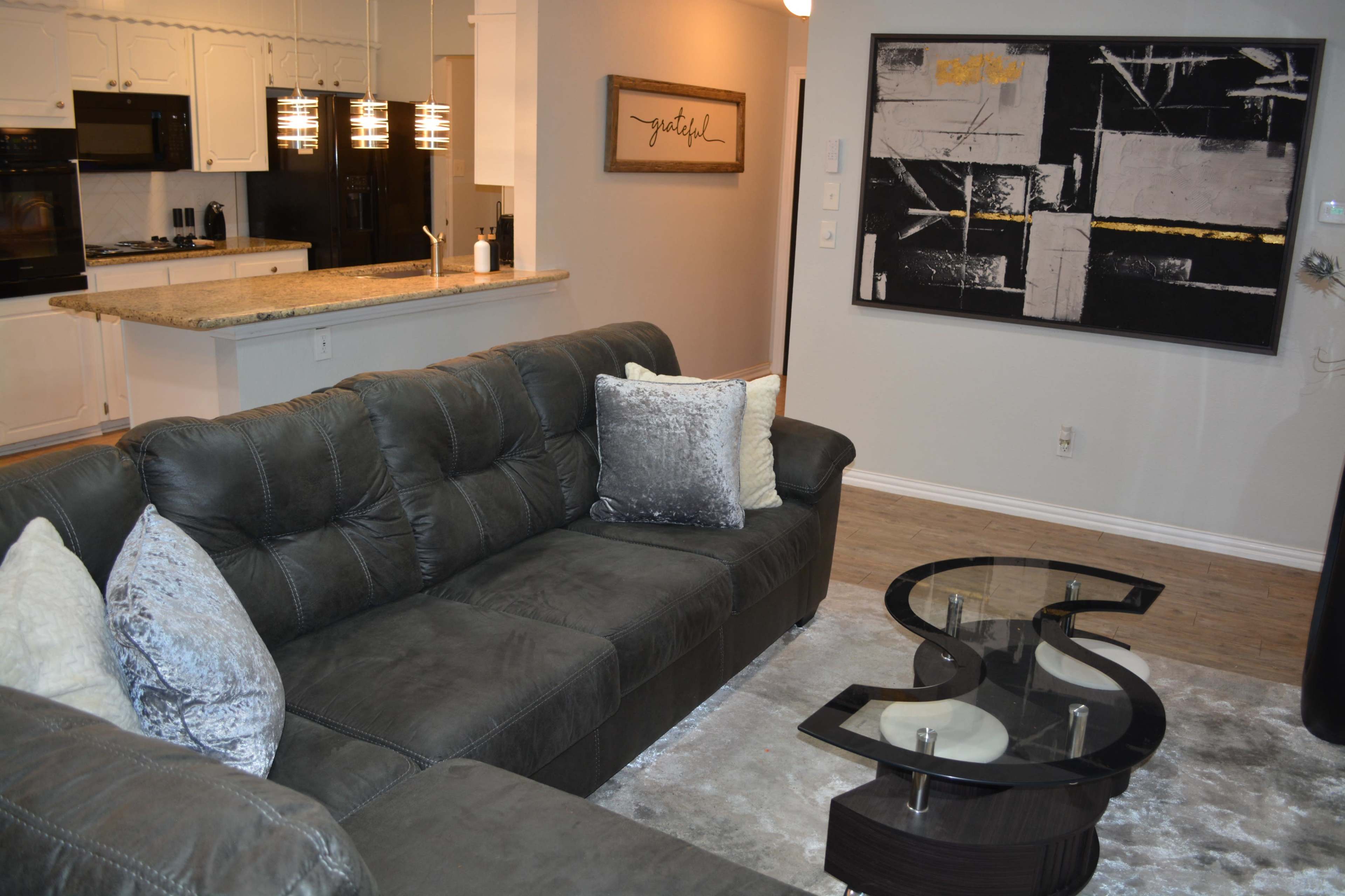 The image shows a modern living room with a dark sectional sofa, a glass coffee table shaped like an "S," and an abstract black and white painting on the wall.