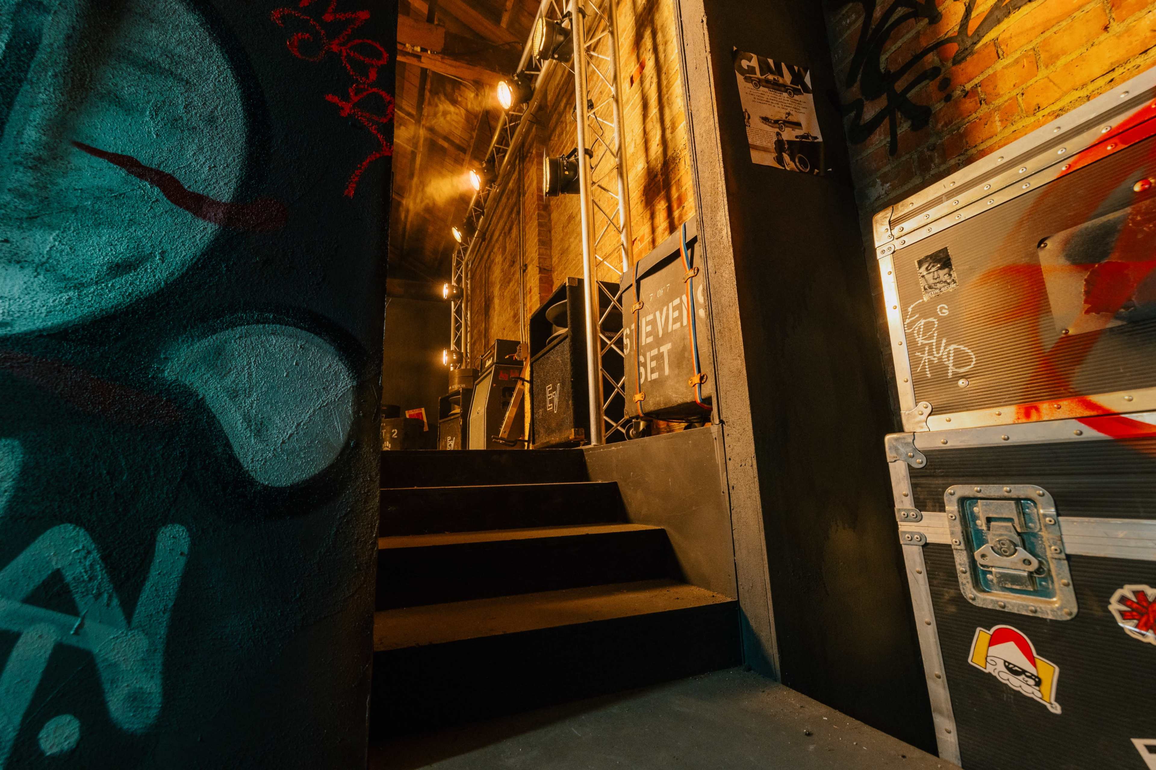 The image shows a dimly lit staircase leading up from a dark hallway, with graffiti on the walls and equipment cases positioned along the sides.