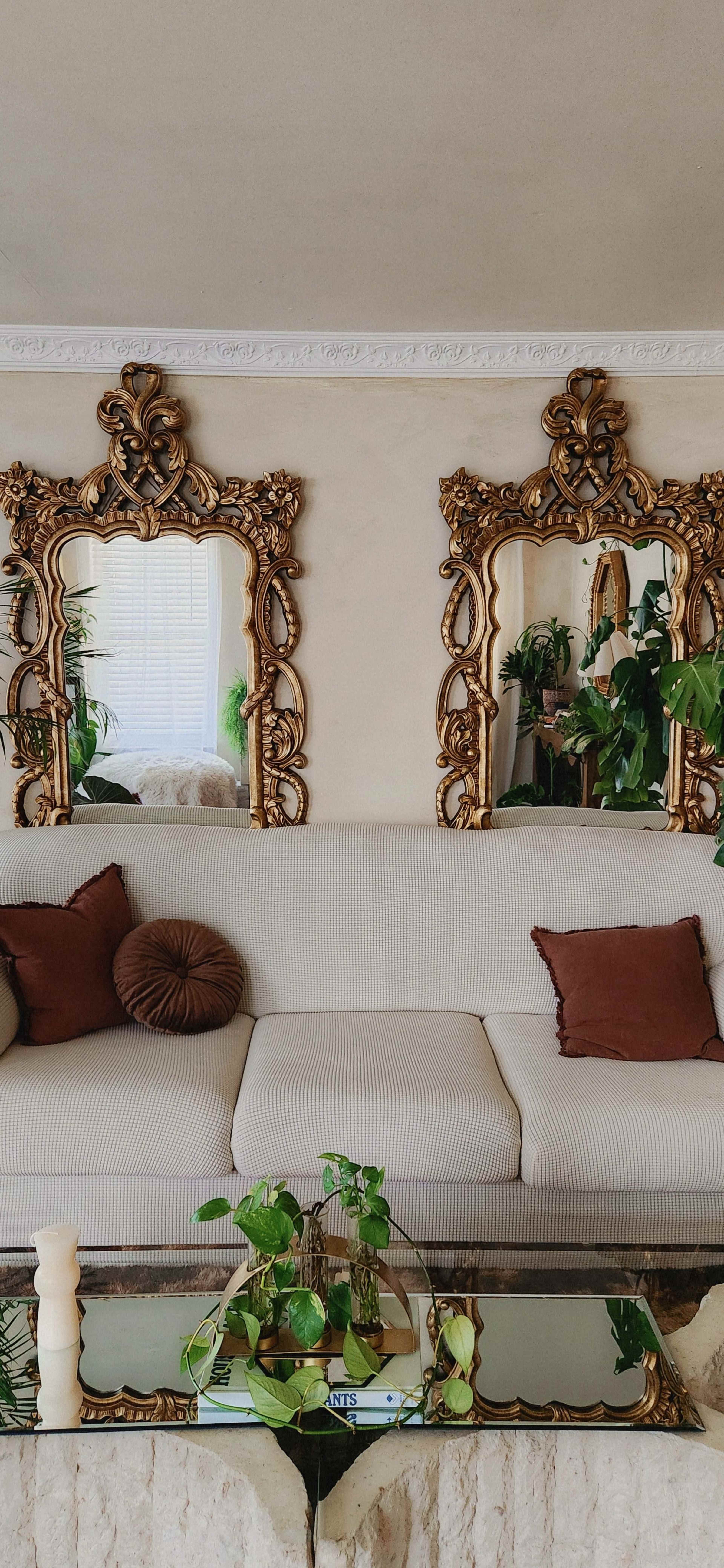 The image shows a living room with a striped beige sofa adorned with brown pillows, flanked by two ornate mirrors and surrounded by indoor plants.