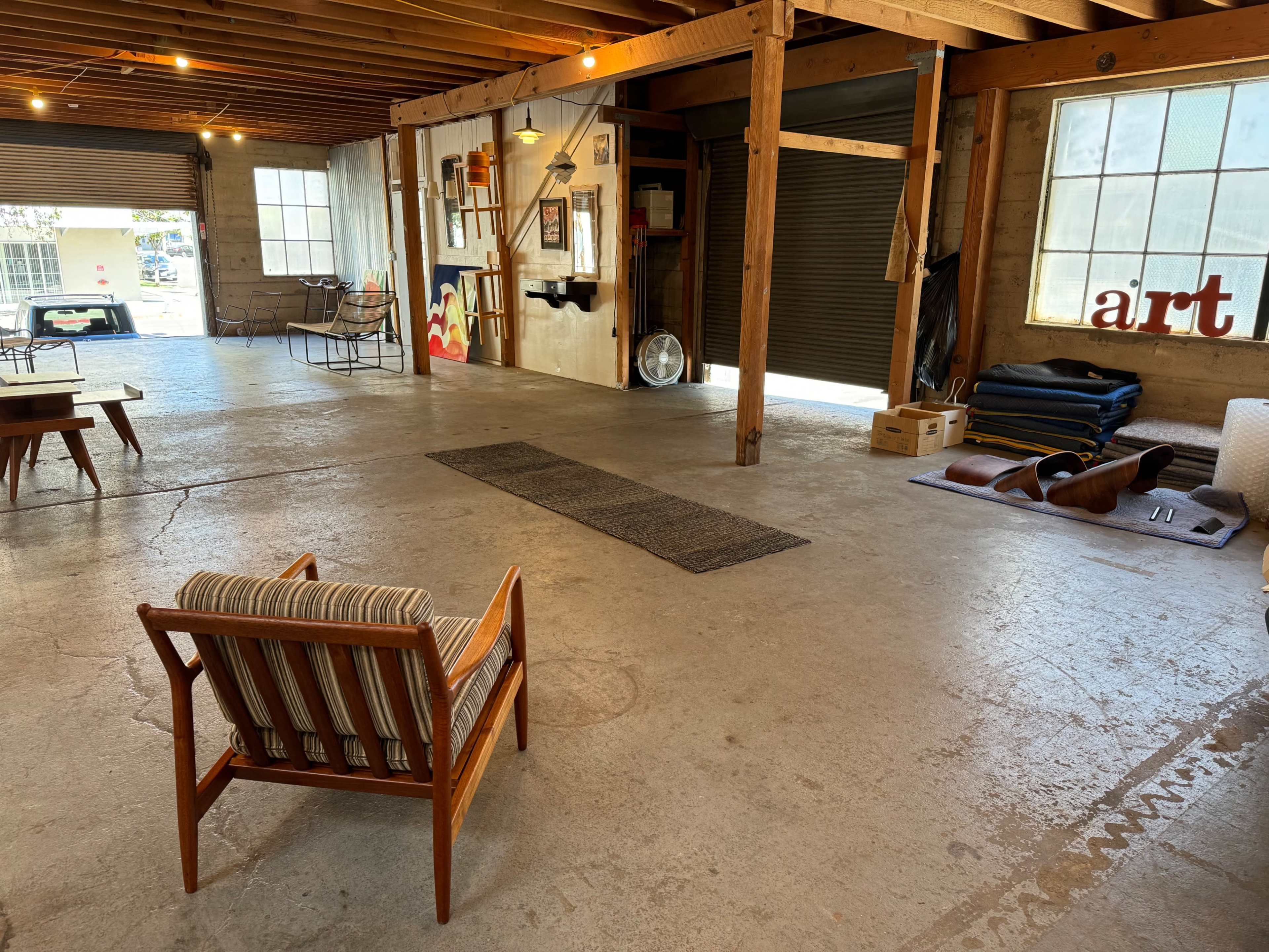 An open warehouse space with concrete flooring, wooden beams, a striped chair, and various furniture items scattered around.