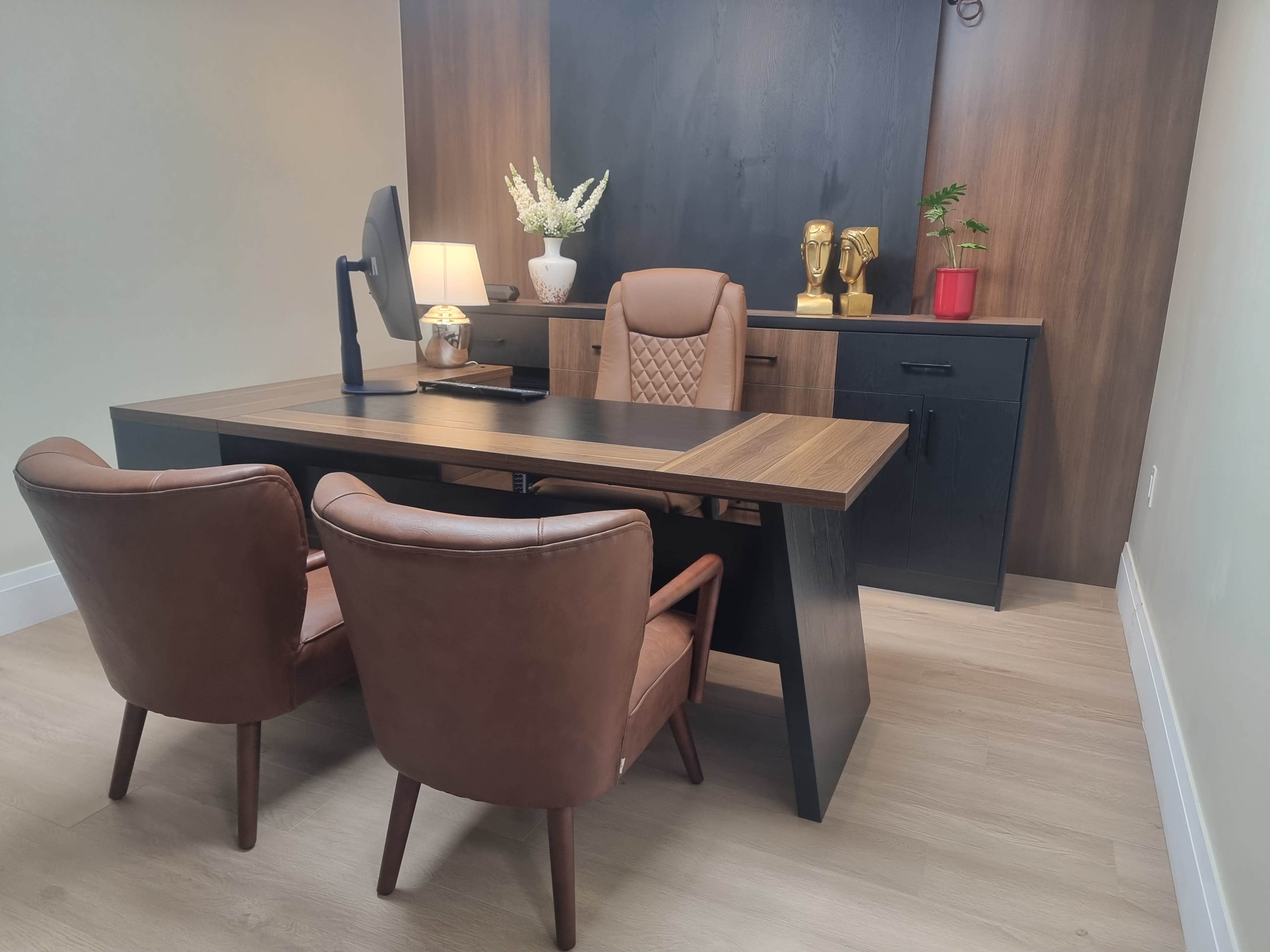 The image shows a modern office space featuring a wooden desk, two brown leather chairs, and a dark backdrop with decorative items.
