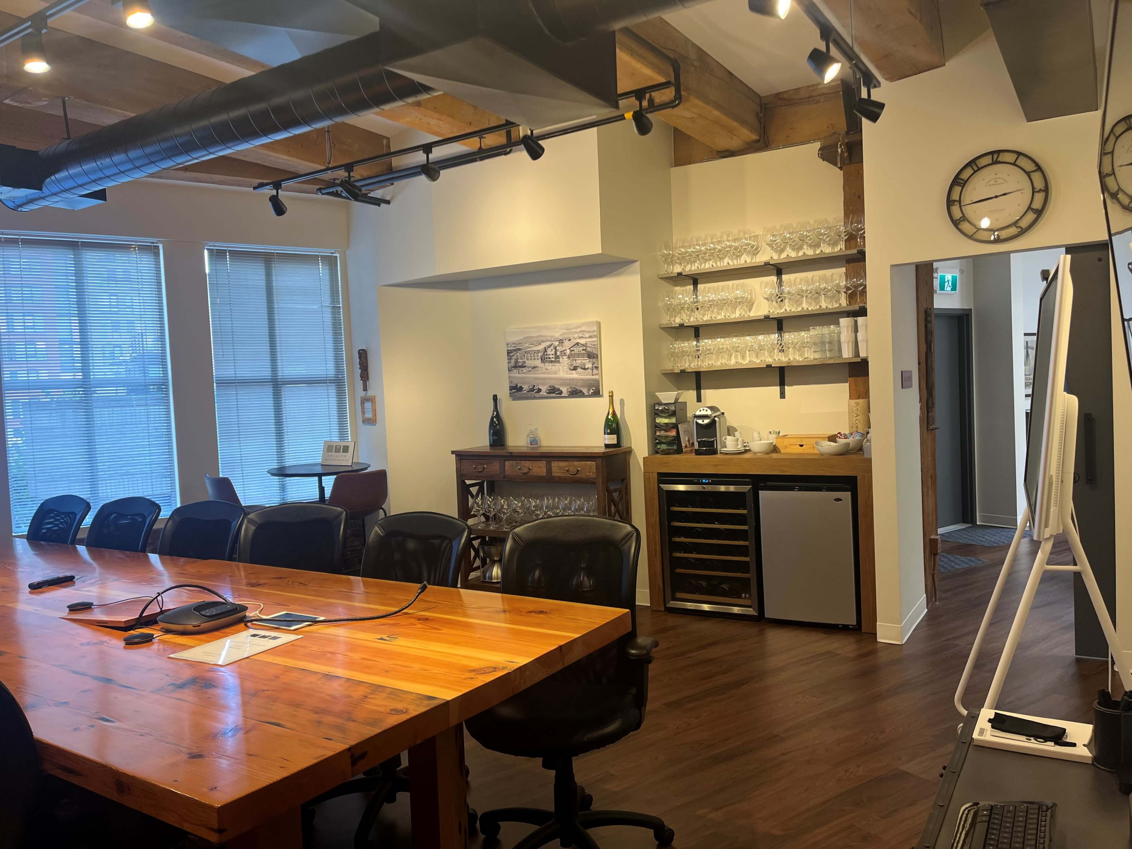 A modern conference room with a large wooden table, black chairs, and a wall-mounted screen, alongside a kitchenette featuring glassware and a beverage fridge.