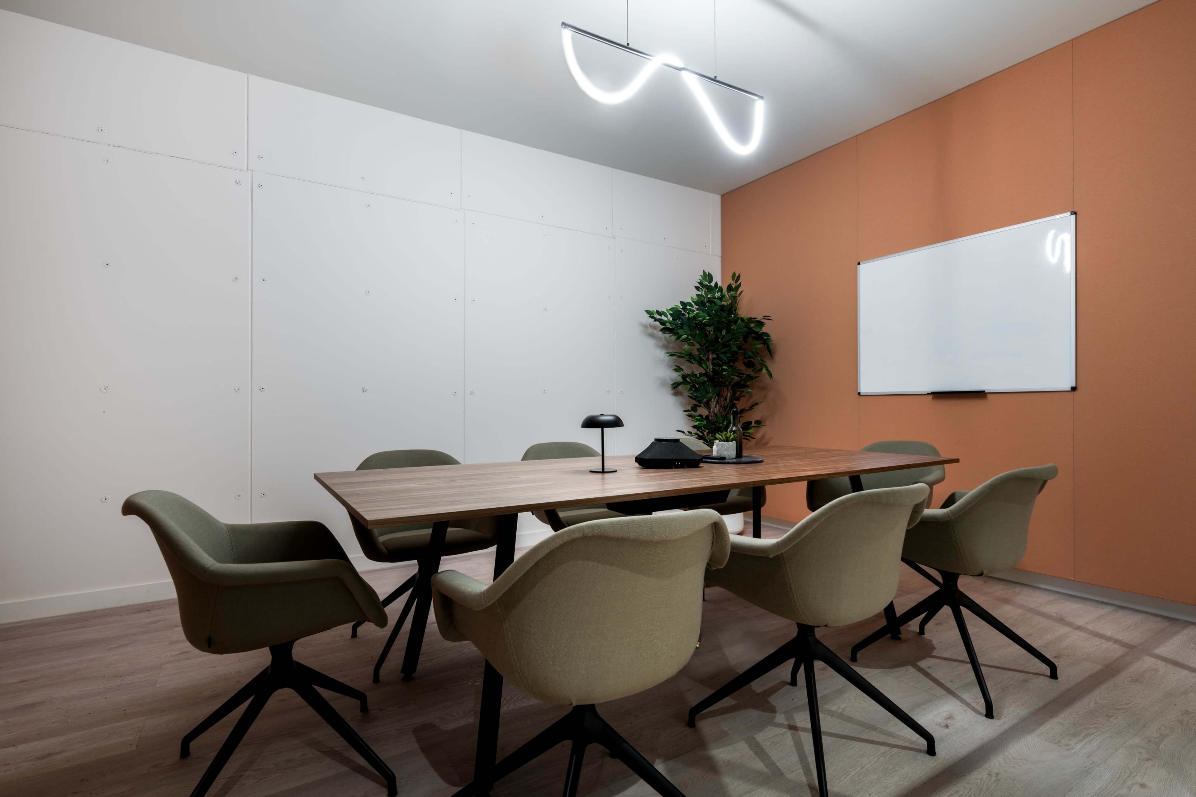 A modern conference room features a large wooden table surrounded by beige chairs, a whiteboard on one wall, and a potted plant in the corner.