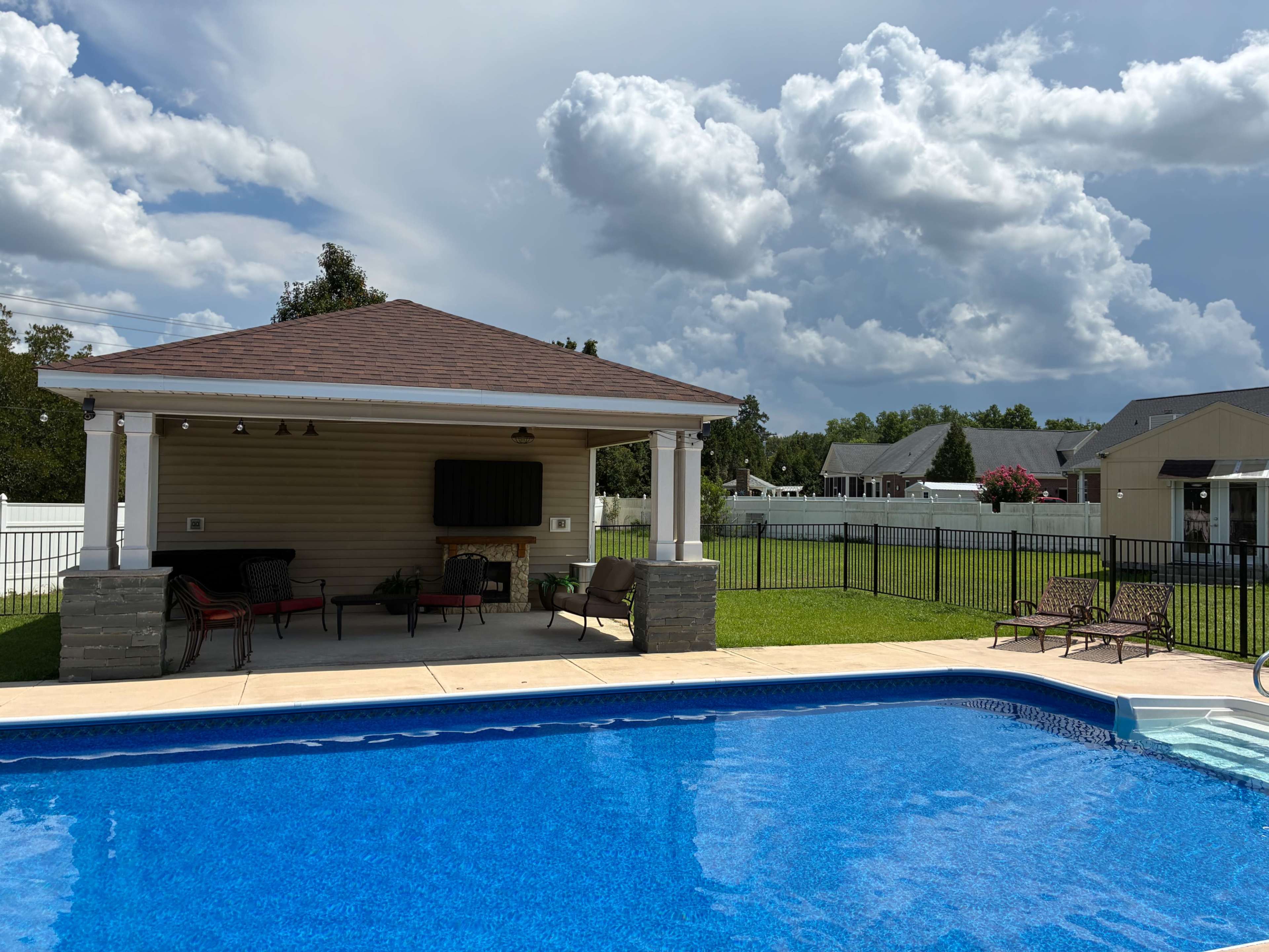 A swimming pool with clear blue water is bordered by a pavilion and several lounge chairs, set against a backdrop of cloudy skies and green grass.