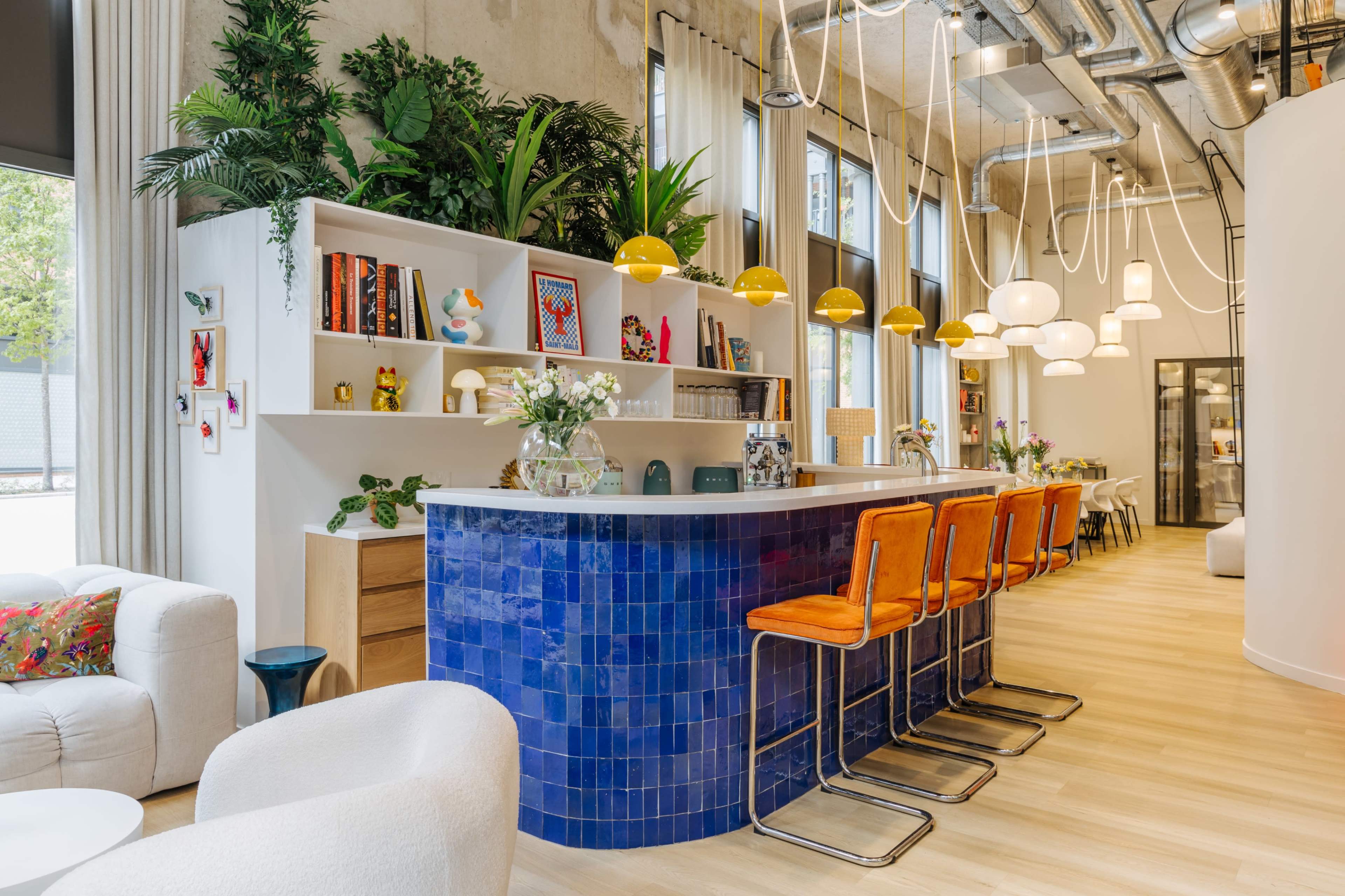 The image shows a modern kitchen space featuring a blue tiled bar with orange stools, surrounded by plants and decorative shelves filled with books and art.