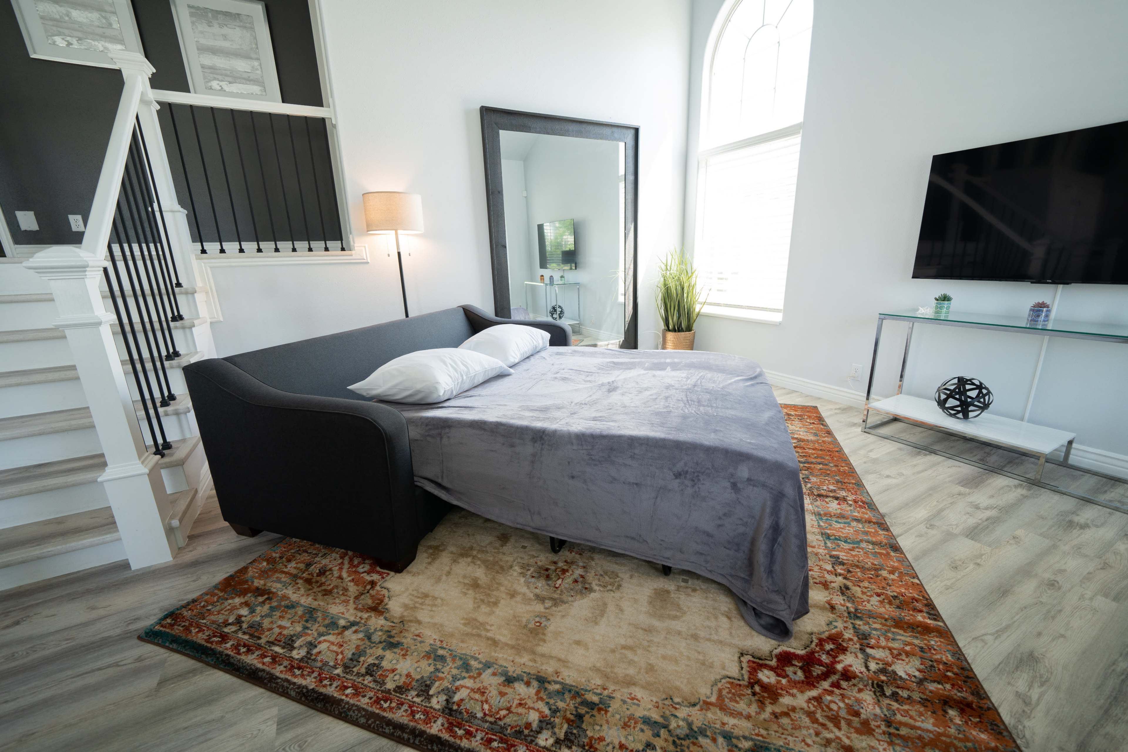 The image shows a modern living space featuring a bed with a gray cover on a decorative rug, a large mirror, a TV on a glass stand, and a staircase.