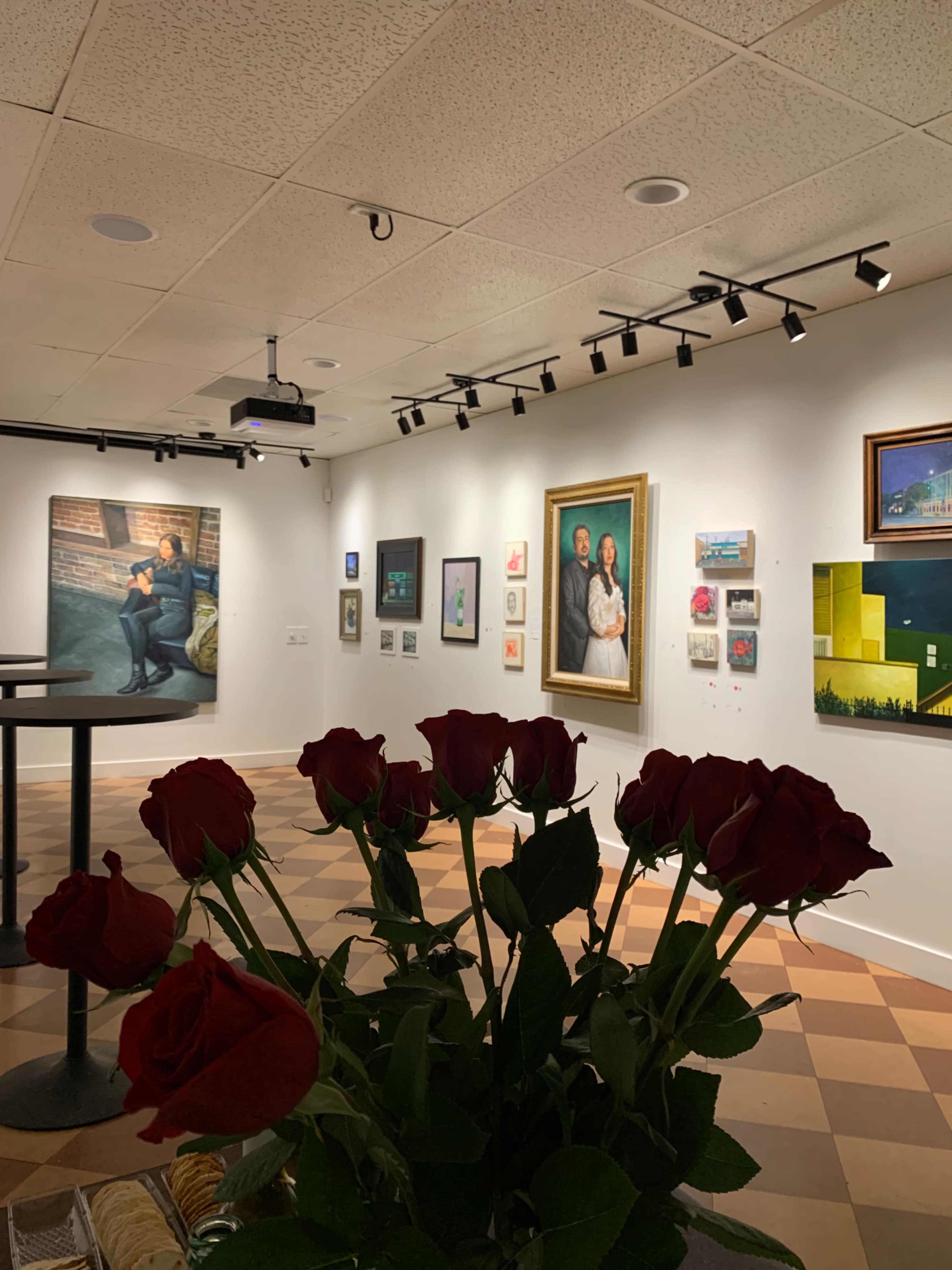 A bouquet of red roses in the foreground contrasts with an art gallery displaying various paintings on white walls.