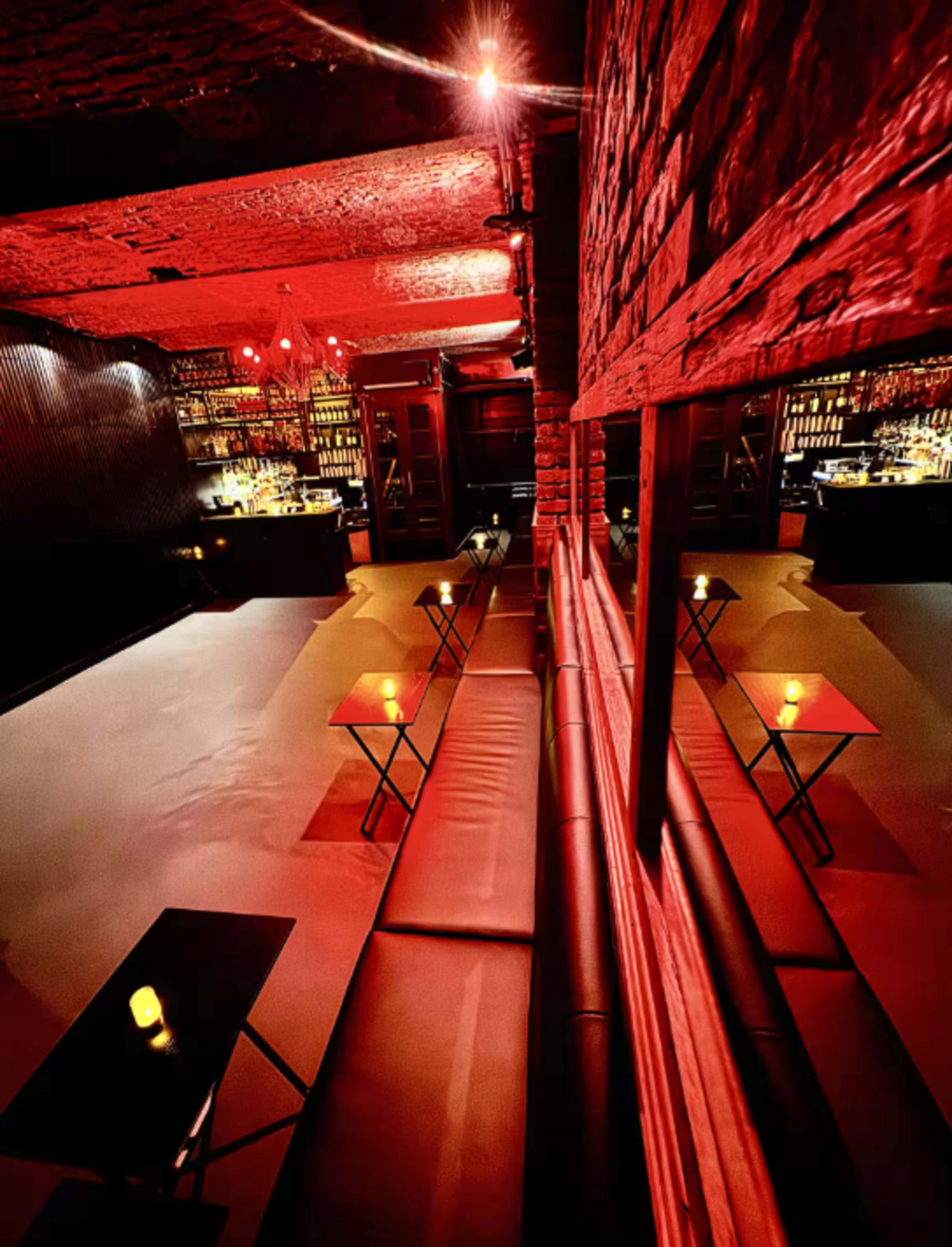 The image shows a dimly lit lounge with red lighting, featuring wooden walls, low tables with candles, and a bar in the background.