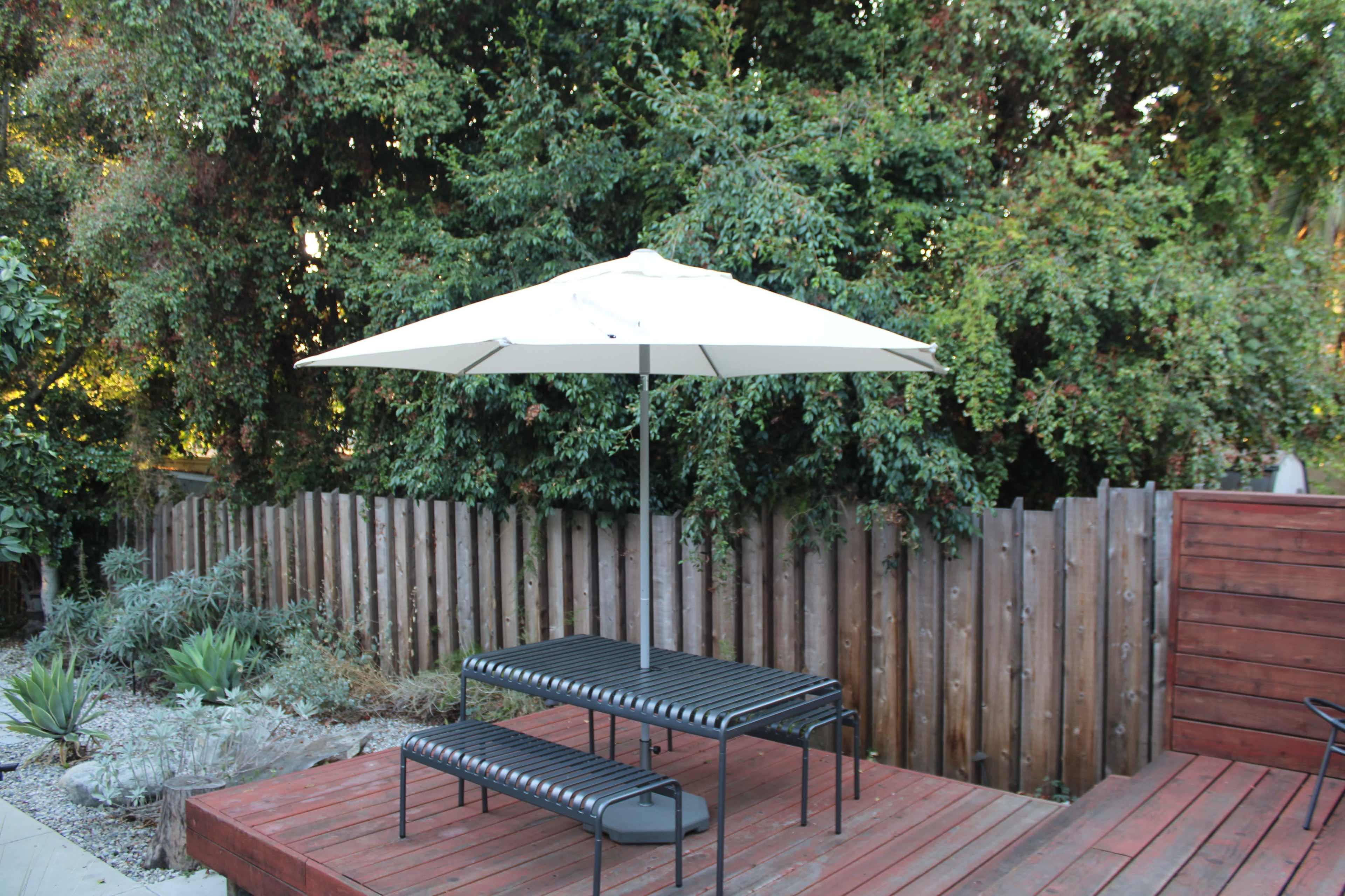 A black metal table with benches sits under a white umbrella on a wooden deck surrounded by a wooden fence and greenery.