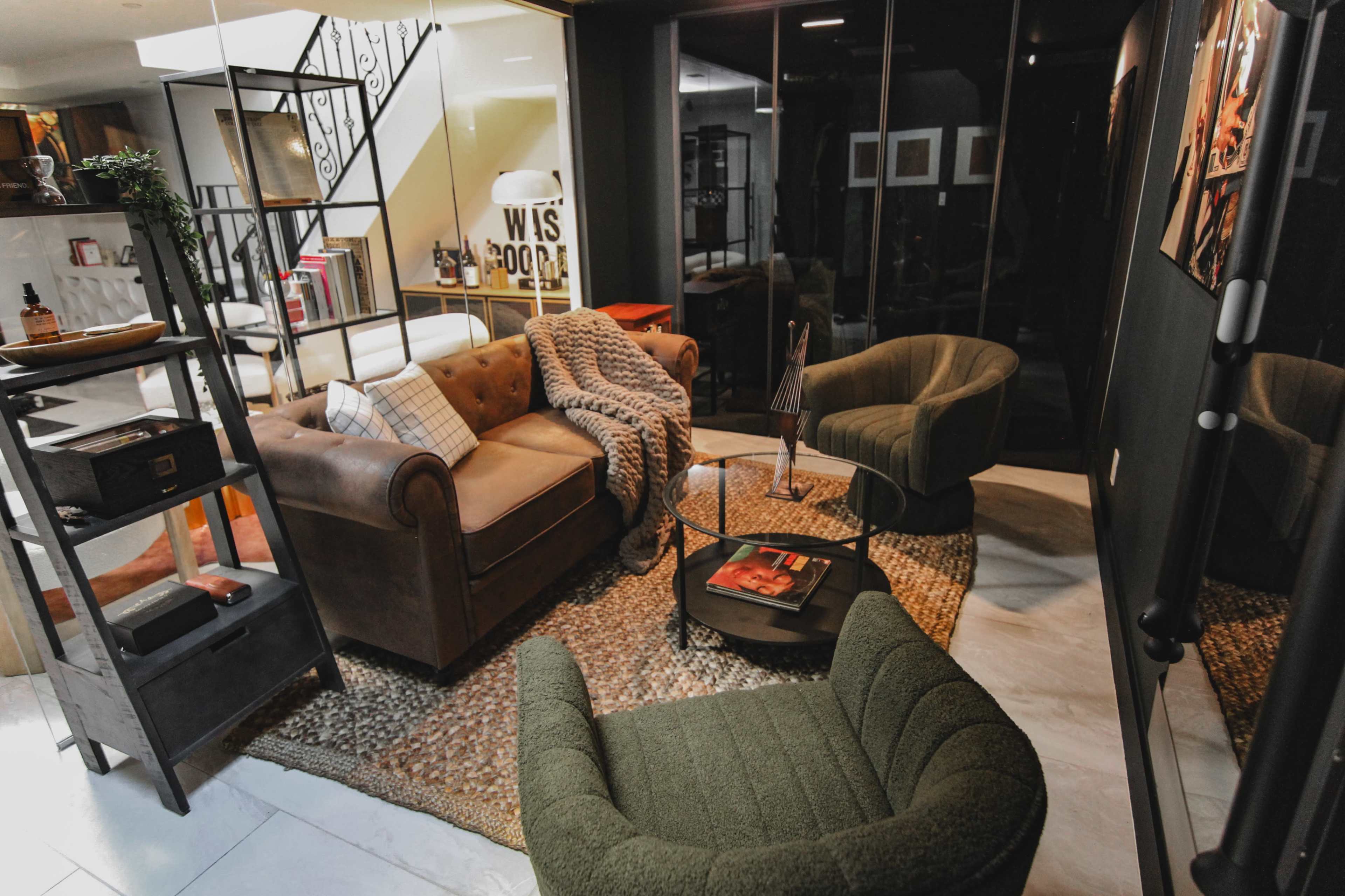 The image shows a cozy living area featuring a brown leather sofa, two green upholstered chairs, a round coffee table, and a decorative rug, all arranged in a modern, well-lit space.