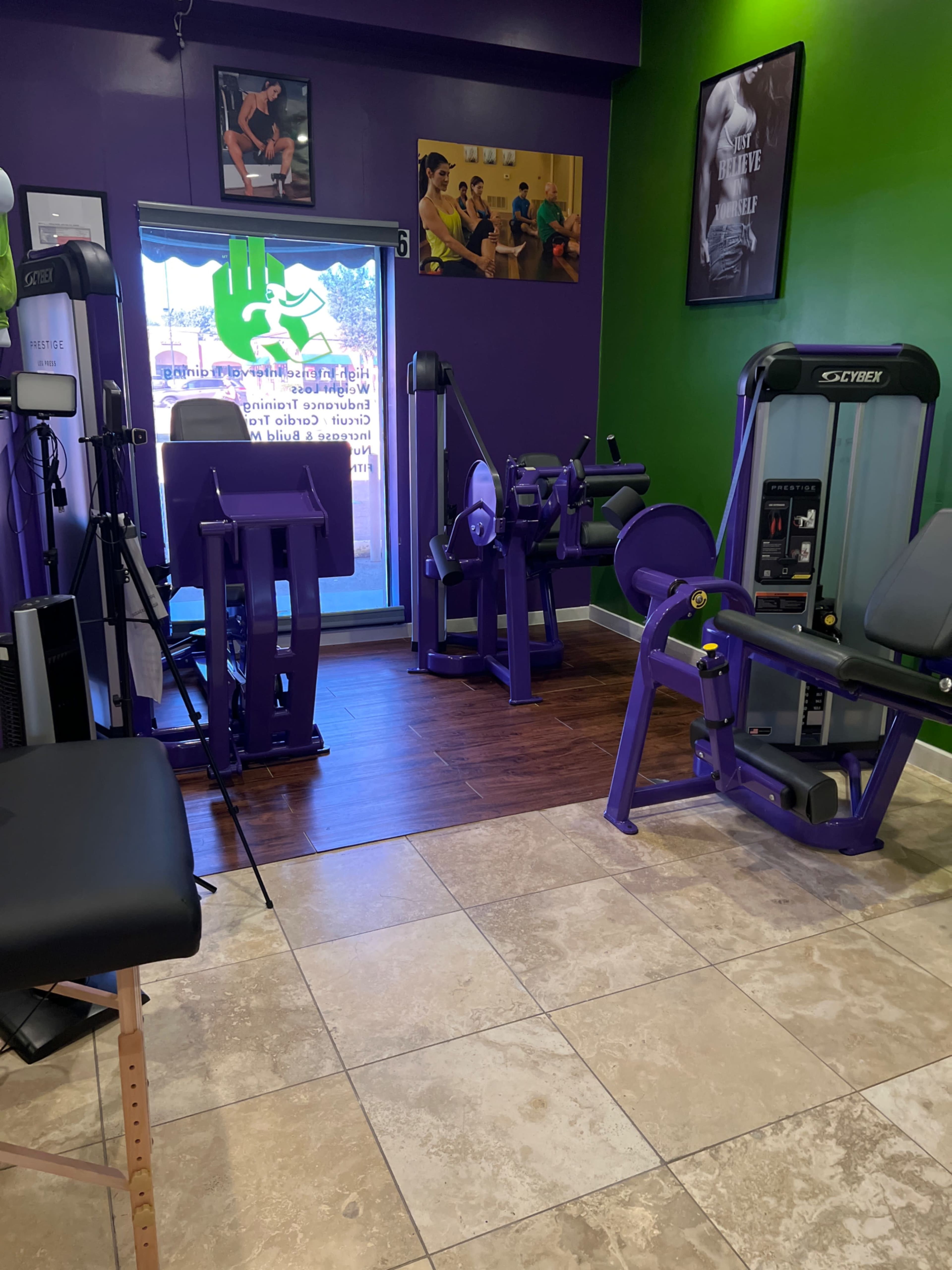 The image shows a fitness studio with several pieces of gym equipment, including a seated leg curl machine and a multi-use strength training machine, set against a vibrant green and purple accent wall.