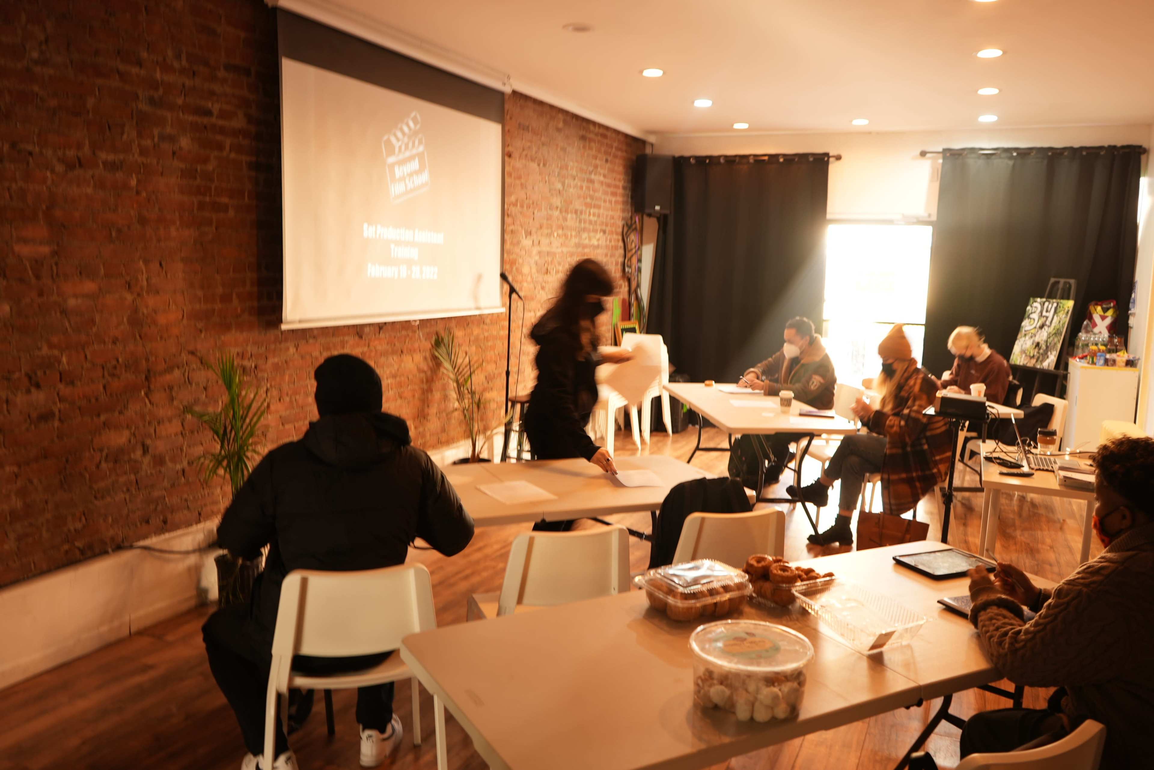 A group of people sits at tables in a well-lit room with exposed brick walls, watching a presentation while some work on laptops.