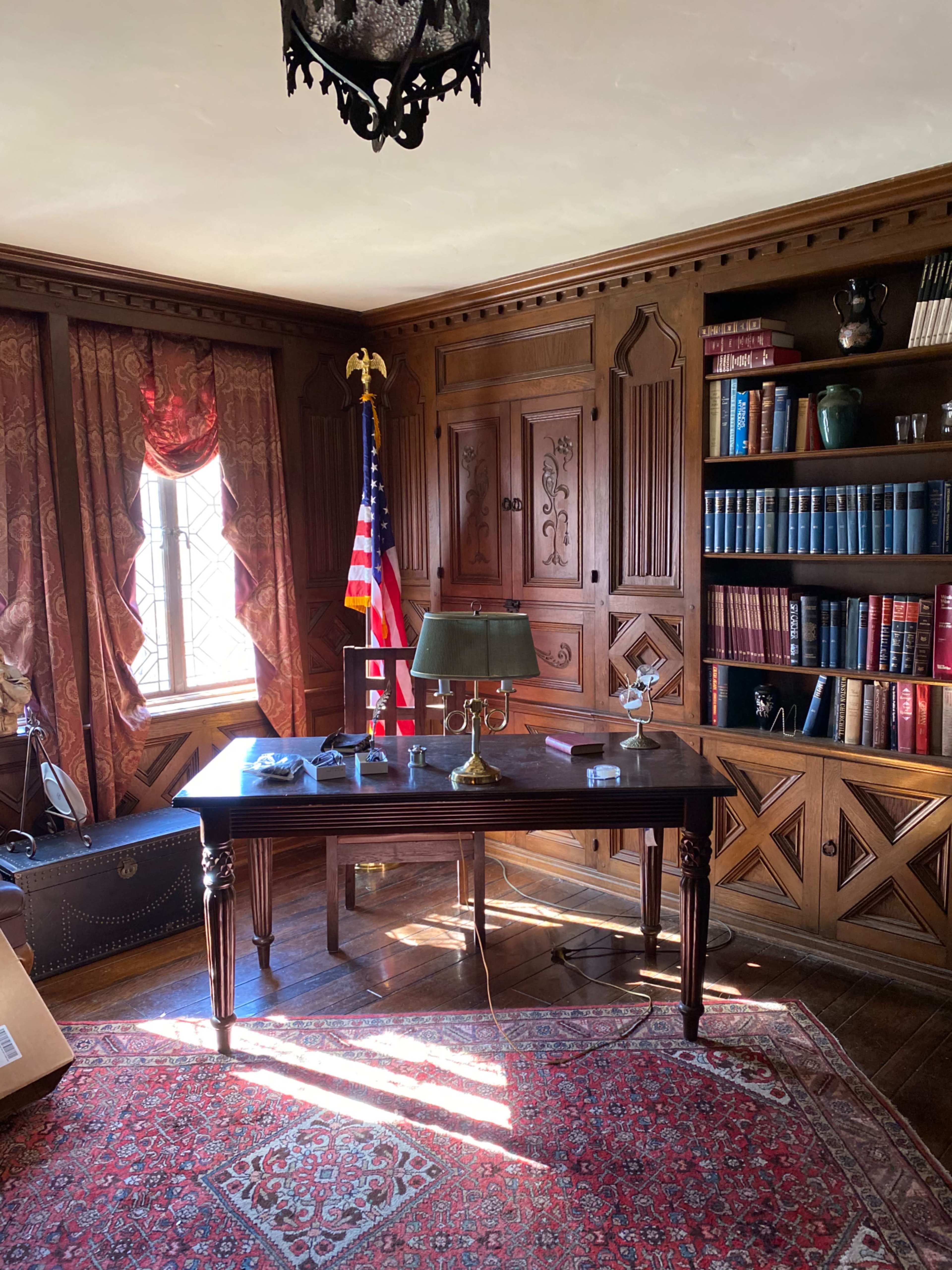 A wooden office furnished with a desk, an American flag, bookshelves filled with books, and a patterned rug on the floor.