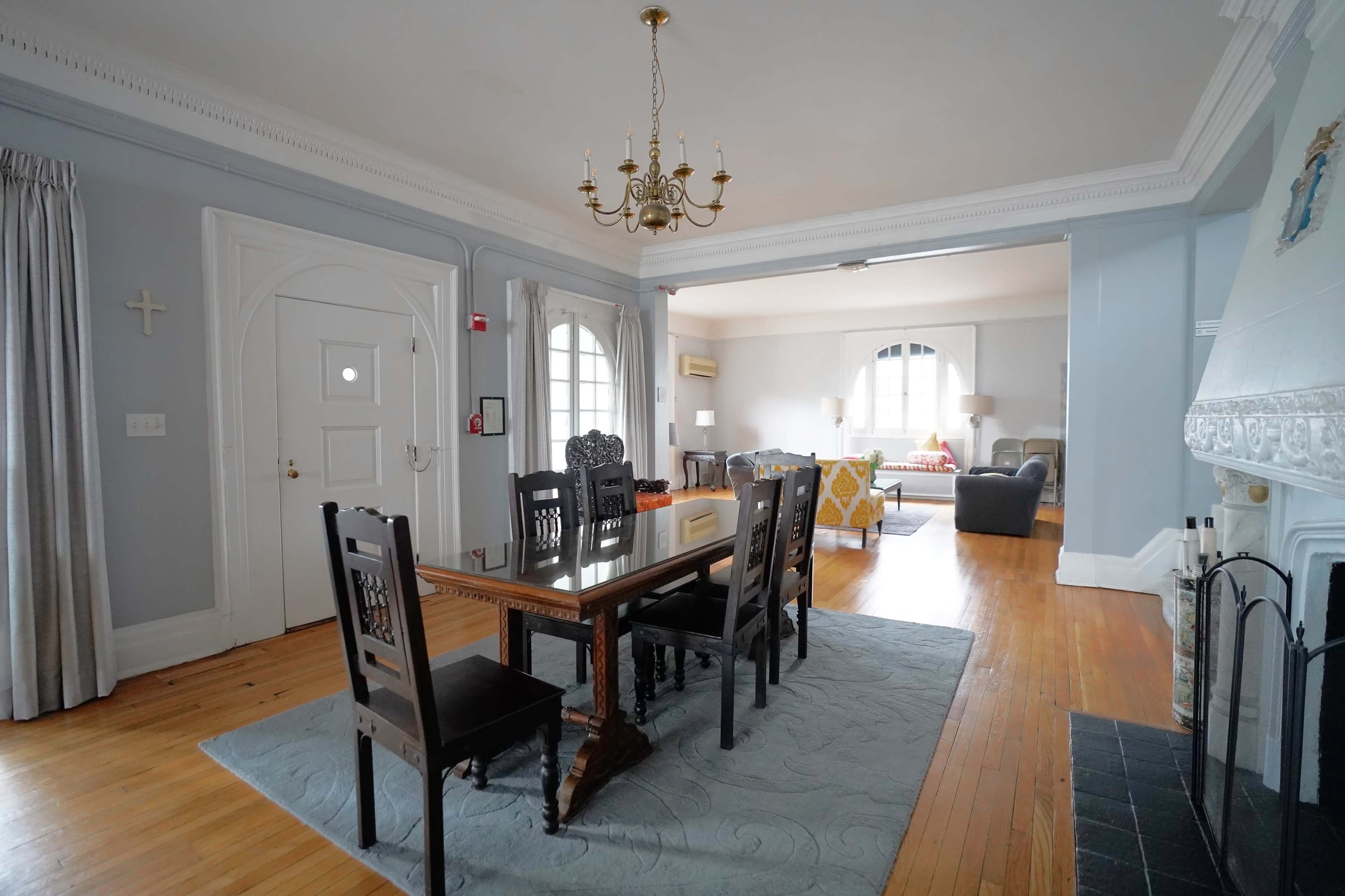 A dining room features a wooden table with black chairs, light gray walls, and a chandelier, leading into a living area with sofas and large windows.