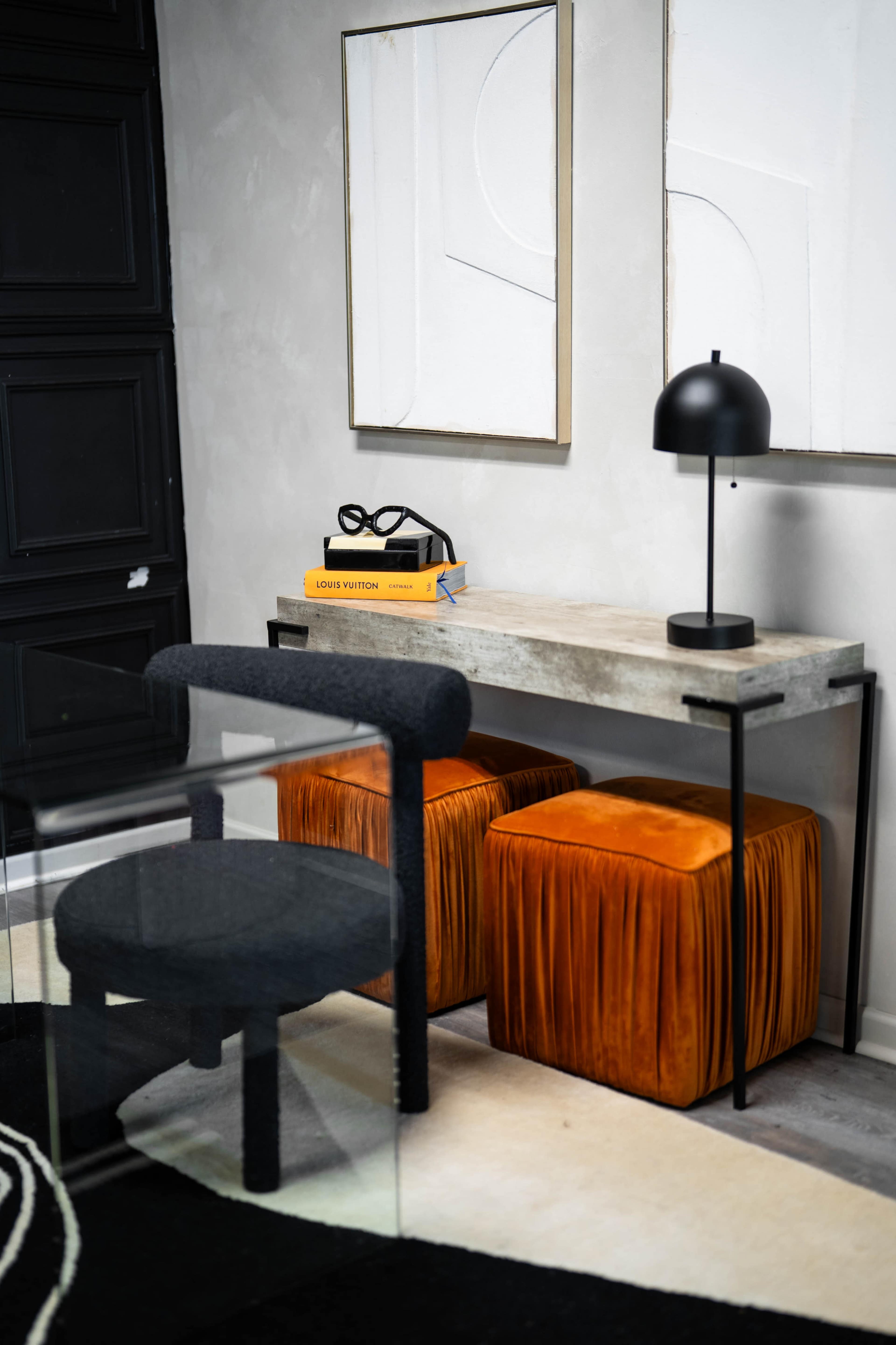 A modern workspace features a concrete console table with orange poufs, a black lamp, and decorative books beside two abstract wall art pieces.