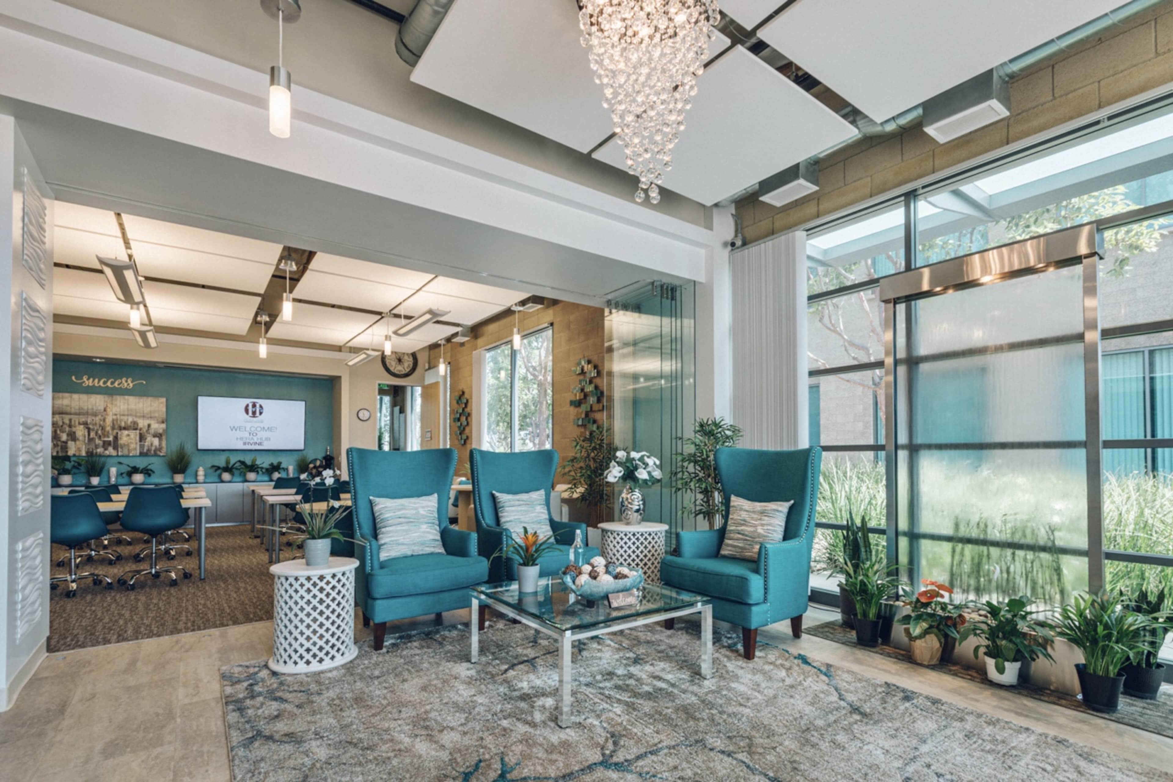 A modern office waiting area features two teal armchairs, a glass coffee table, and a backdrop of plants and a conference room.