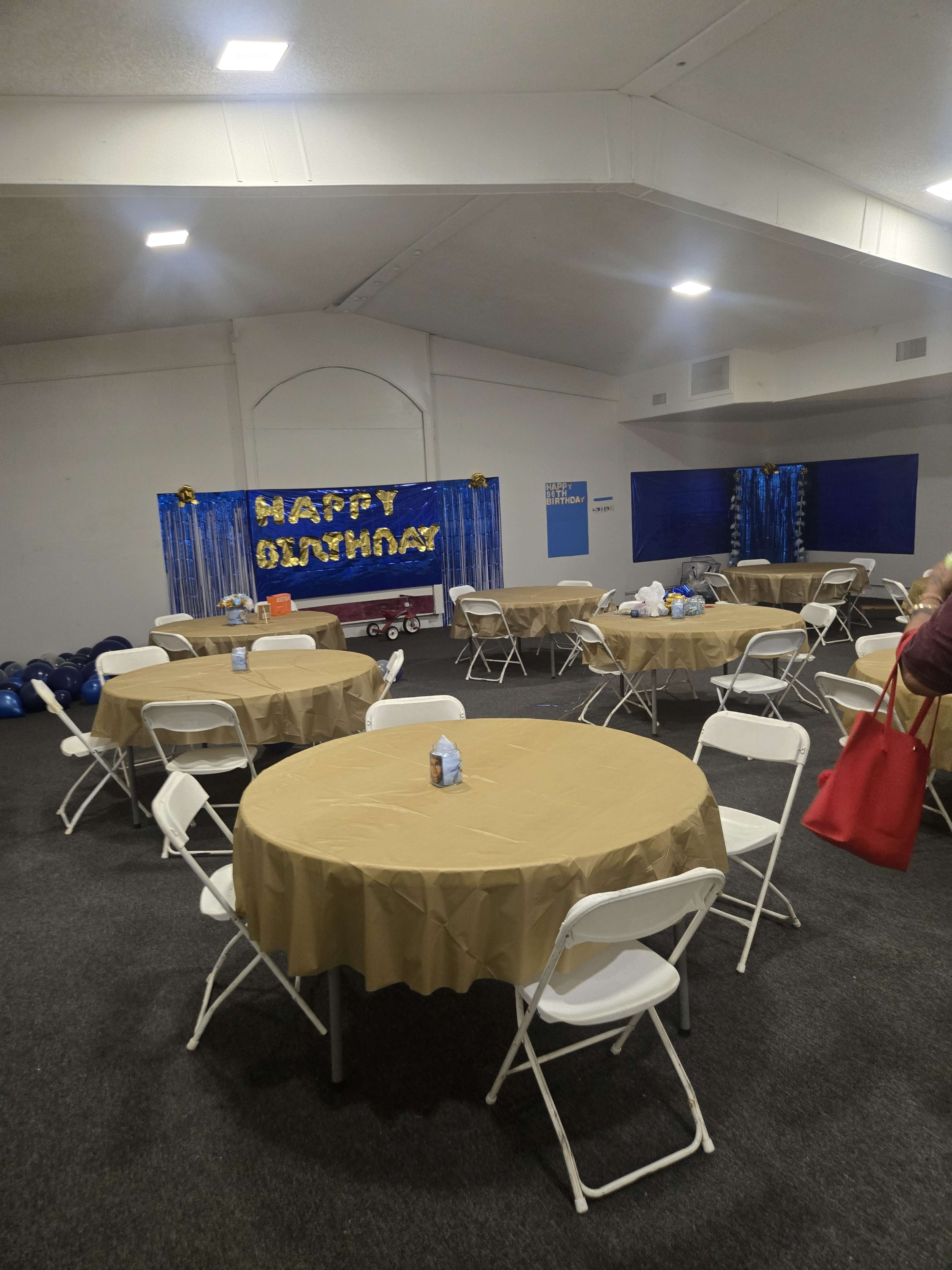 The image shows a spacious indoor setup for a birthday celebration, featuring several round tables with gold tablecloths, white folding chairs, and a "Happy Birthday" banner in the background.