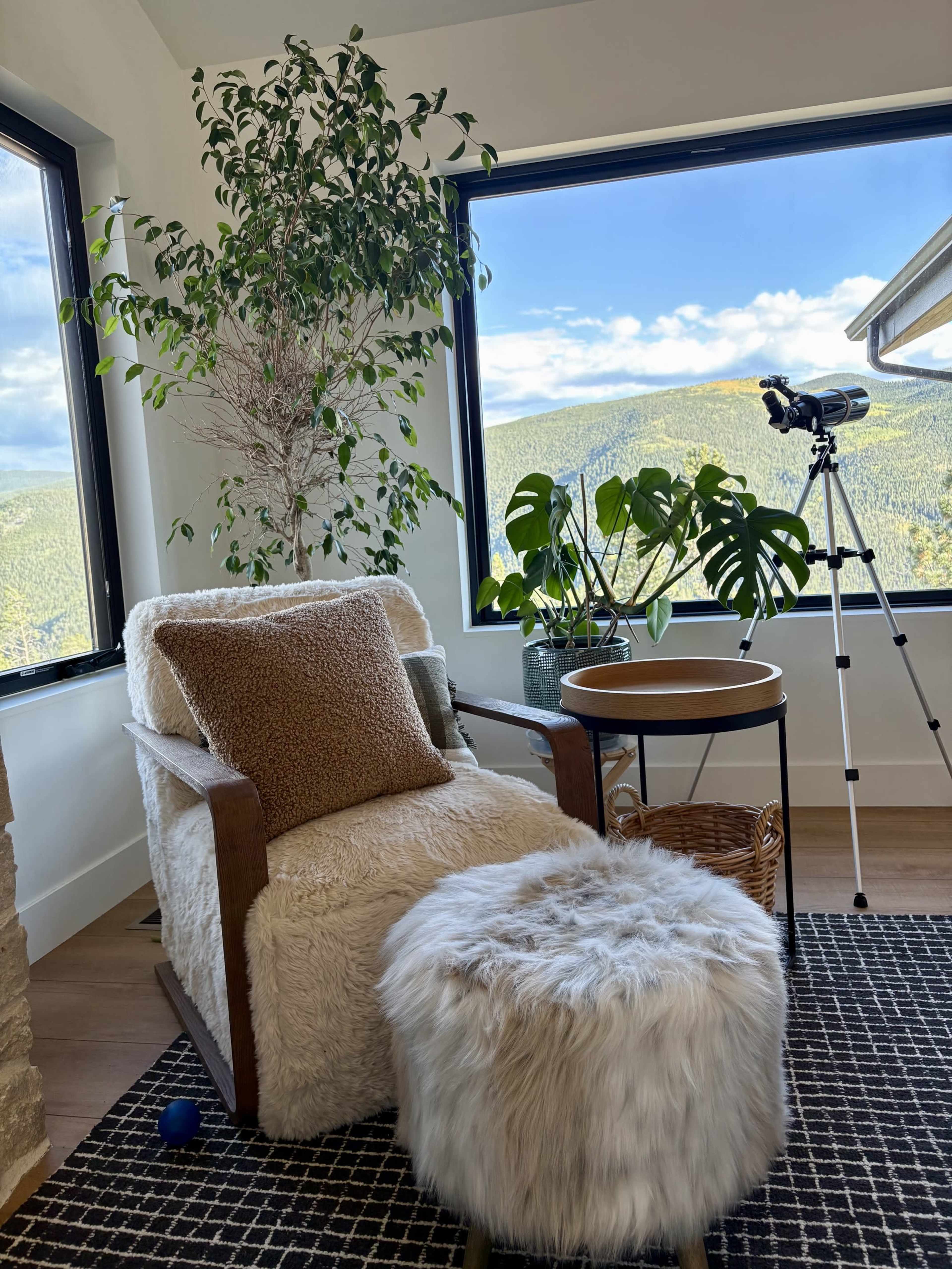 A cozy reading corner features a plush chair, a small side table, and a fluffy ottoman, with large windows showcasing a mountain view and a telescope nearby.