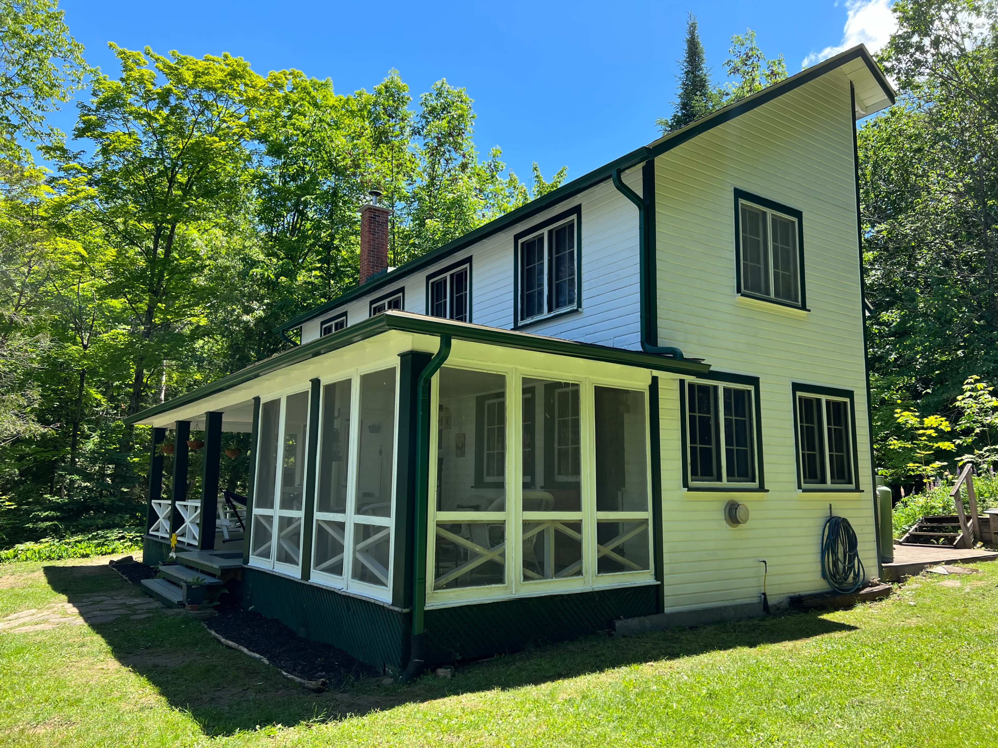 A two-story white house with green trim features a screened porch and is surrounded by lush trees in a rural setting.