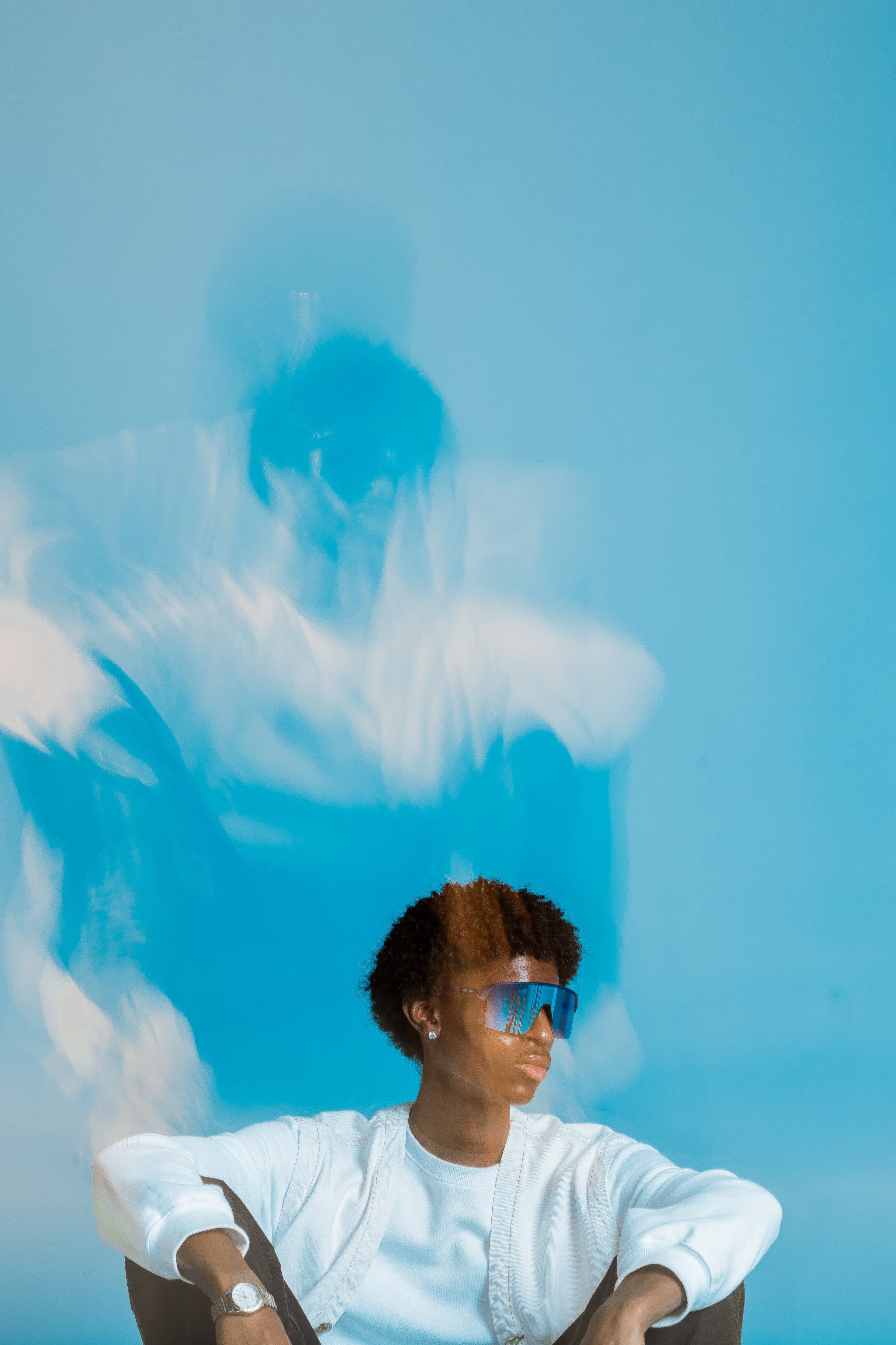 A person wearing sunglasses sits against a blue background while a blurred figure appears to move above them.
