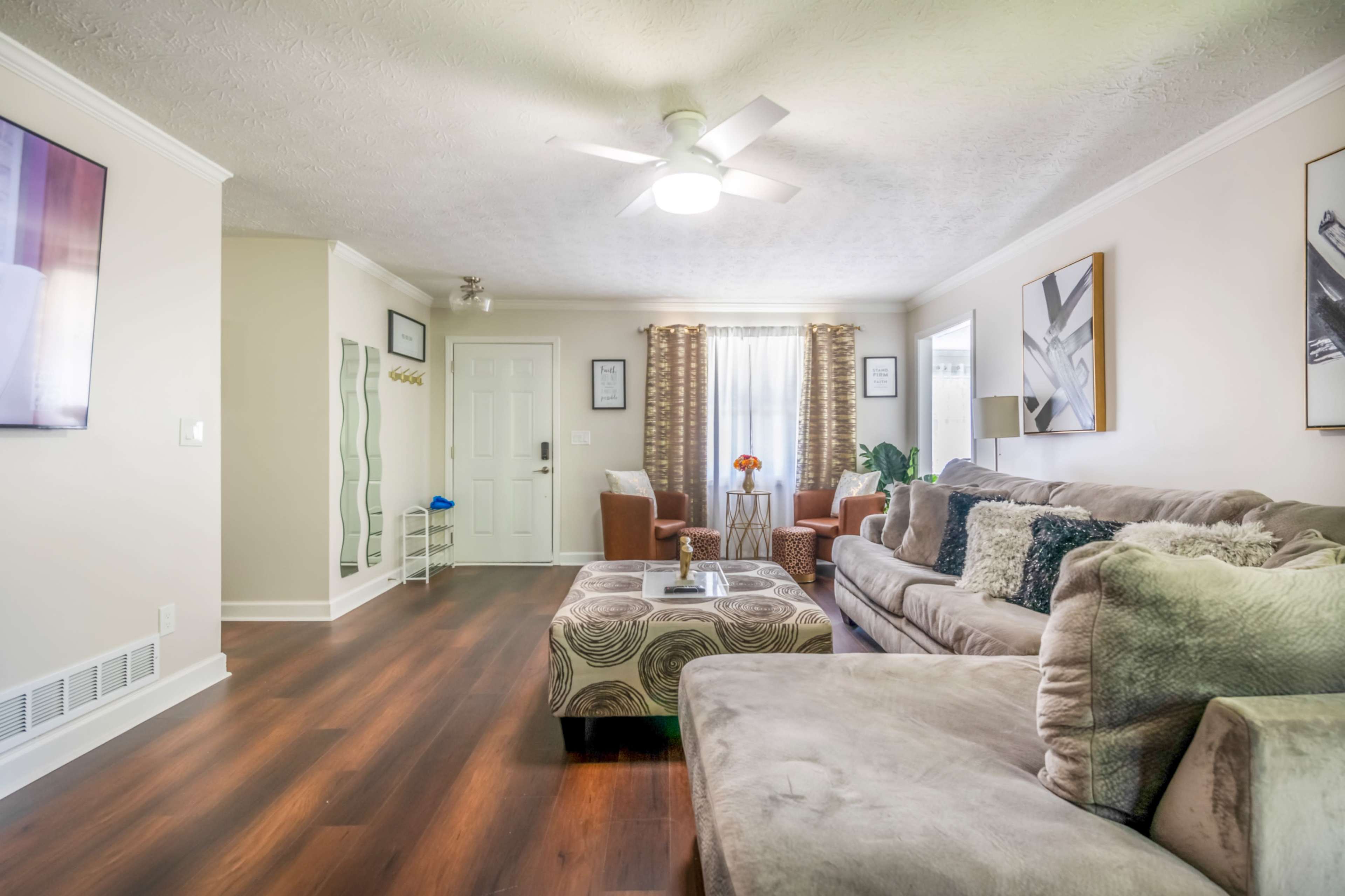 The living room features a beige sofa with decorative cushions, a coffee table, and two armchairs near a window, with a ceiling fan and a television mounted on the wall.