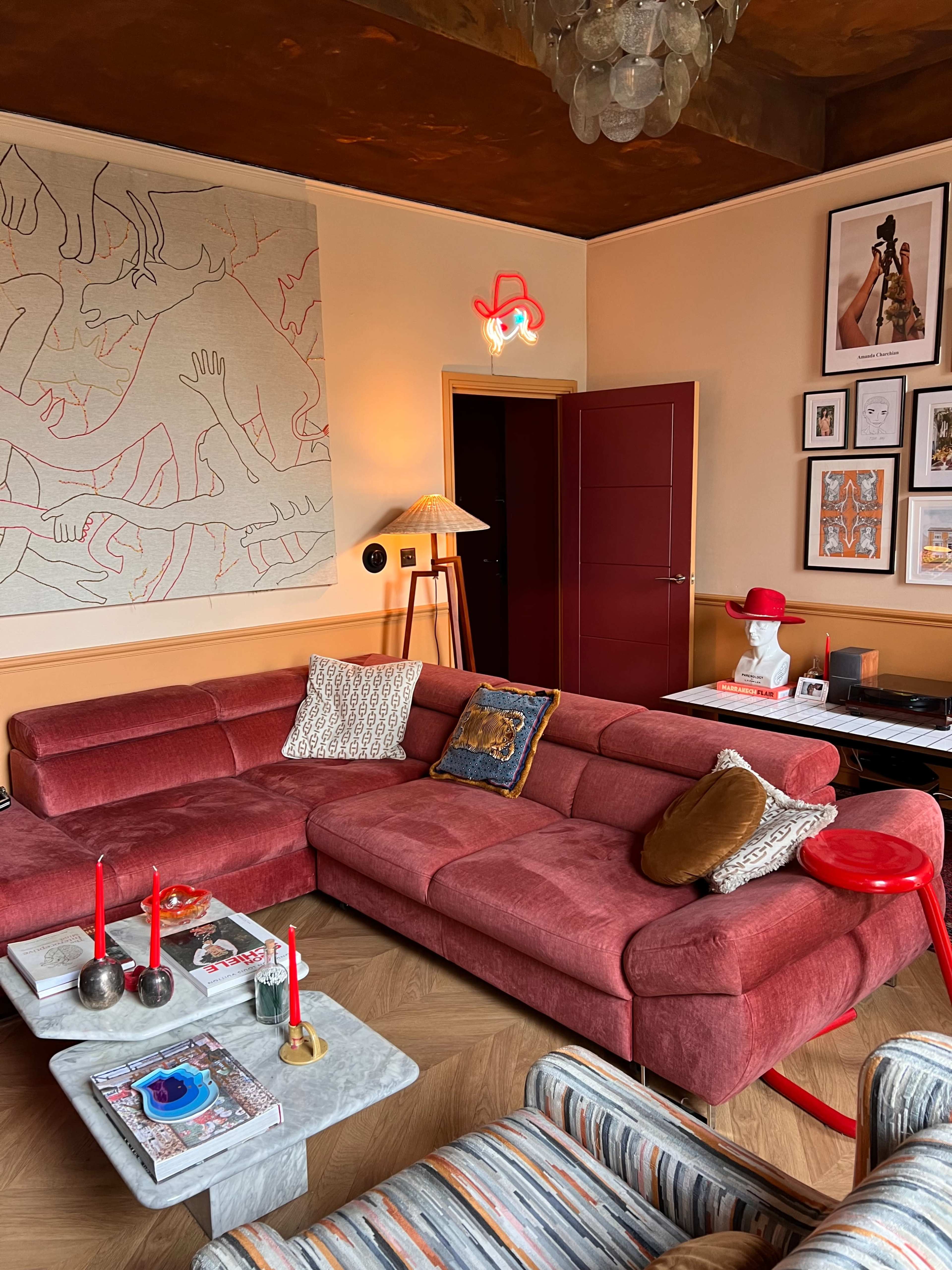The living room features a pink sectional sofa with decorative pillows, a red accent table, and various artworks on the walls.