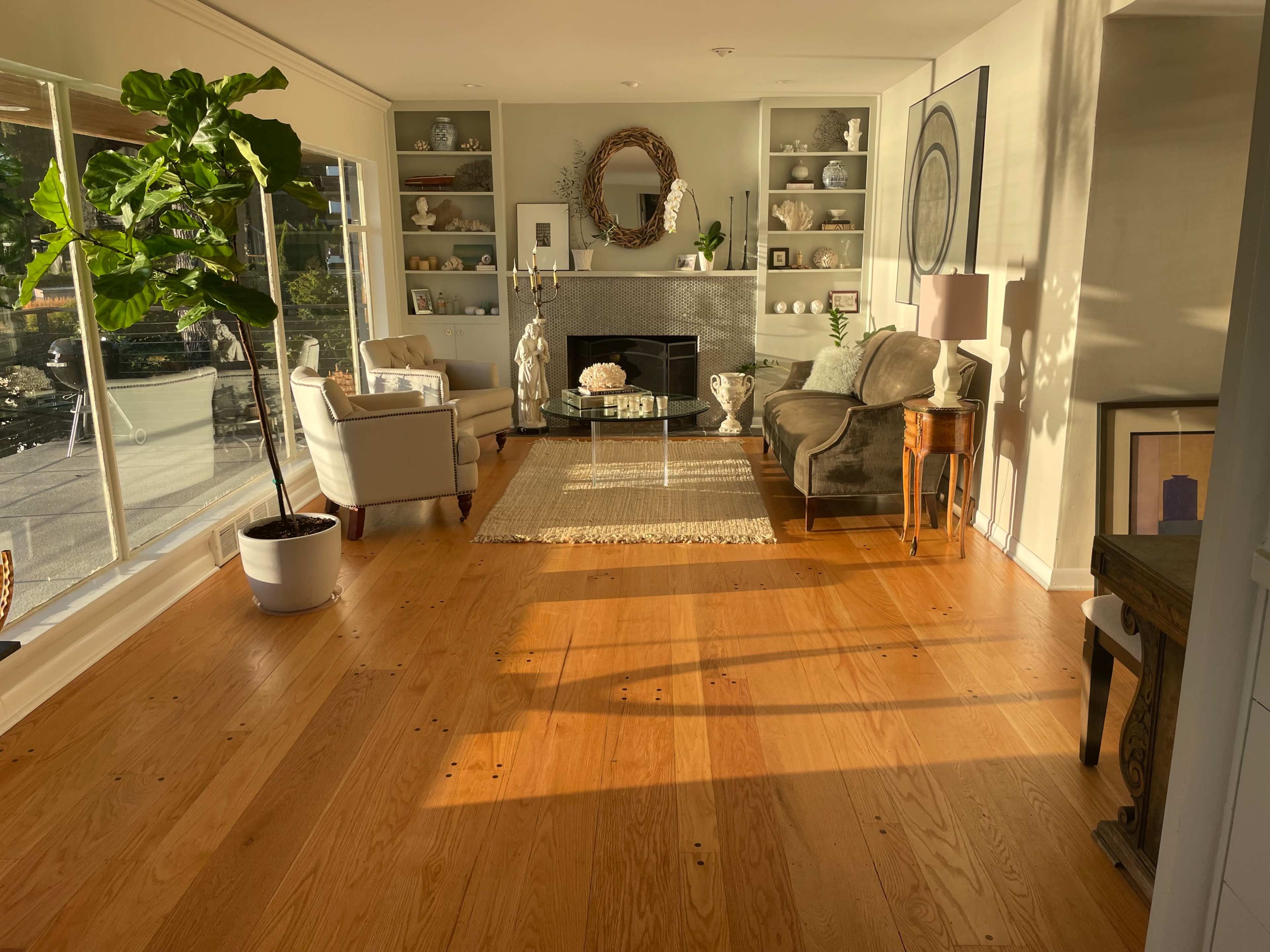 A bright living room features hardwood floors, a large window allowing natural light, a fireplace, and comfortable seating arrangements.