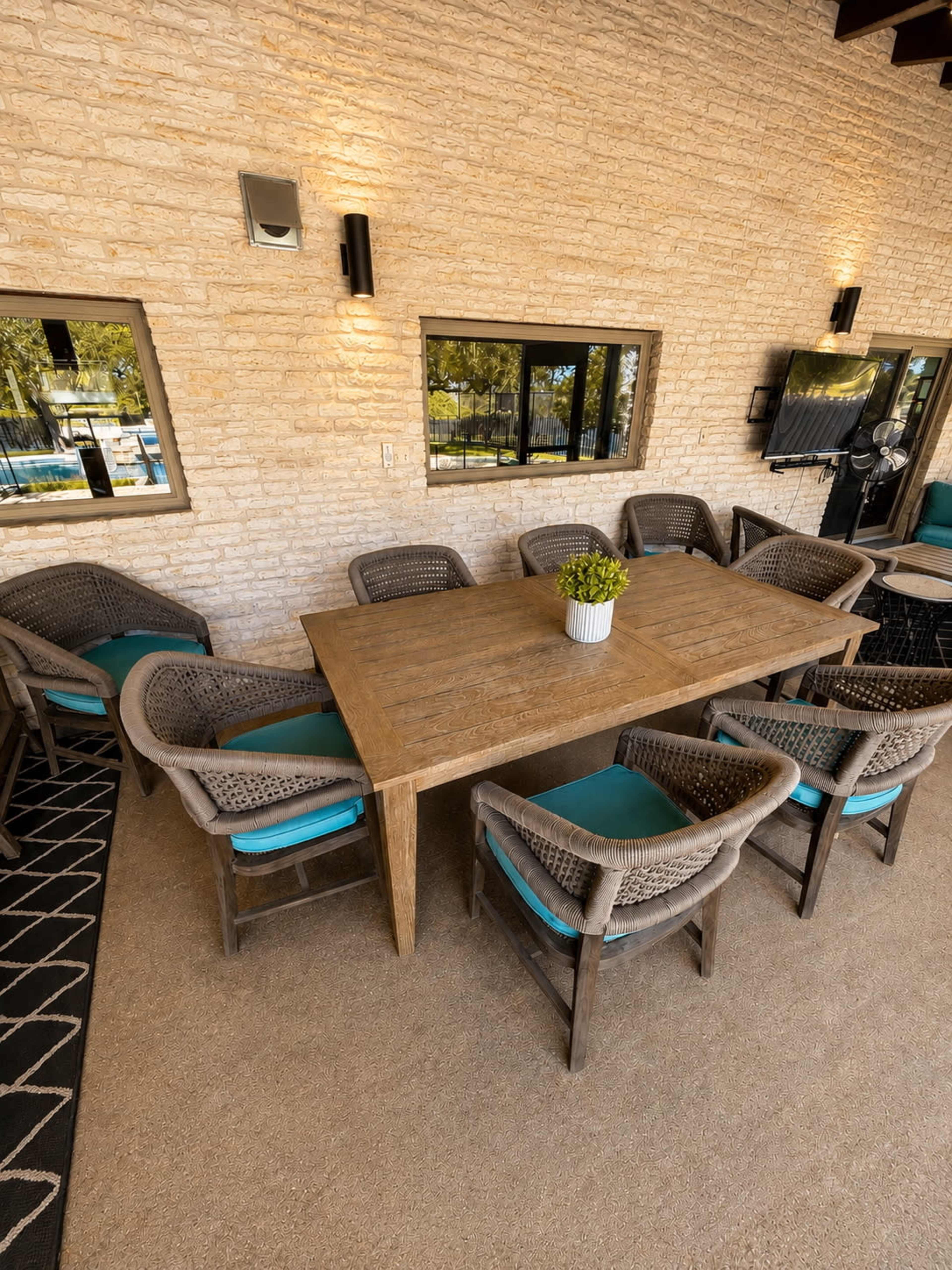 A wooden dining table with eight chairs surrounds it in a well-lit outdoor seating area, accompanied by a decorative plant in a vase.