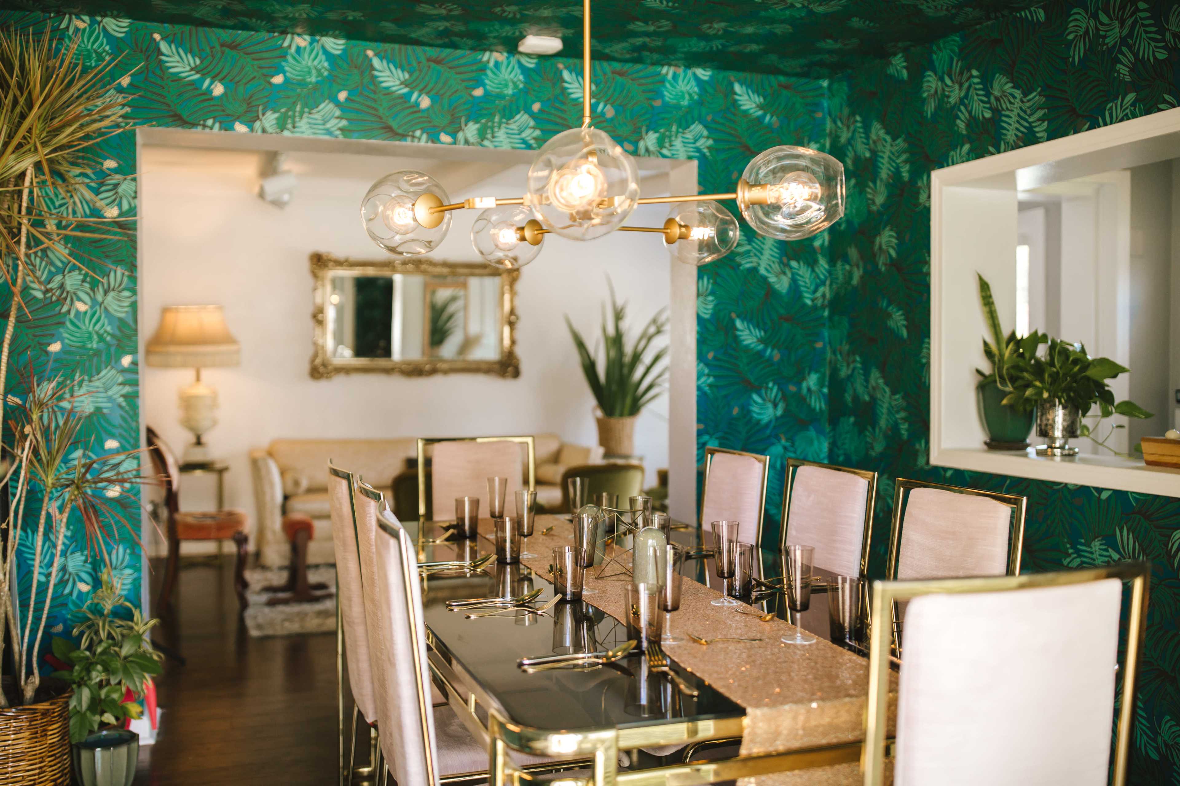 A dining area with a large black table set for a meal, surrounded by gold-framed chairs and decorated with green leaf-patterned wallpaper and potted plants.
