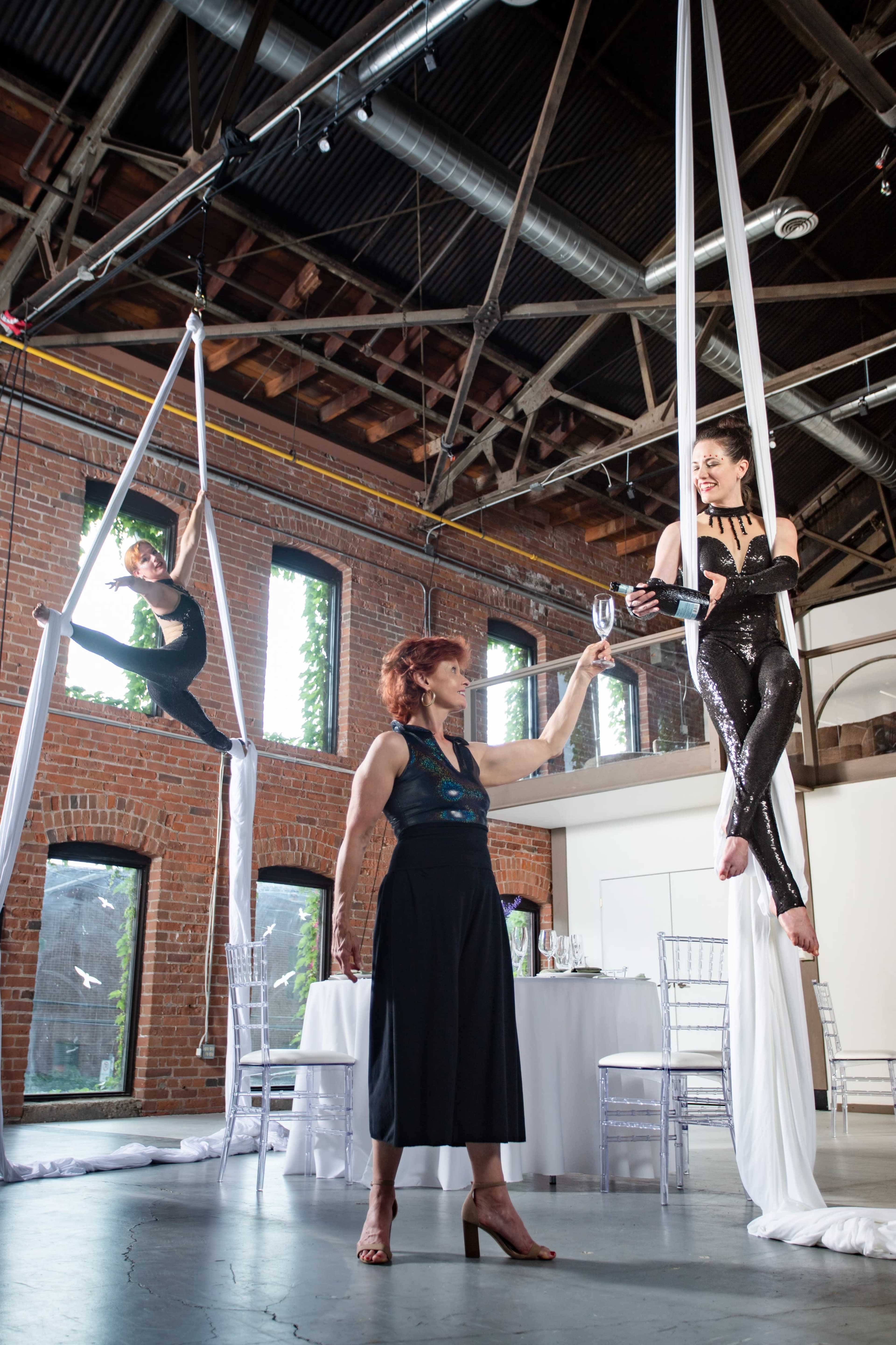 Aerial performers showcase their skills in a spacious, industrial-style venue while a woman in formal attire hands a glass to a performer suspended from silk.