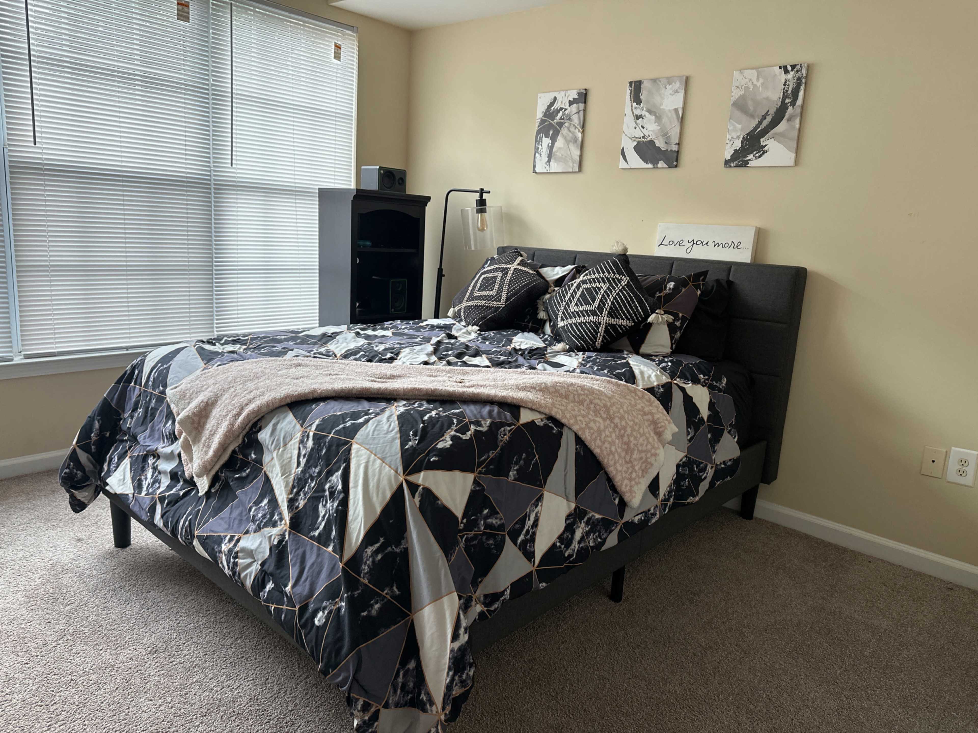 A neatly arranged bedroom with a bed featuring a patterned black and white quilt, a bedside lamp, and three framed pictures on the wall.