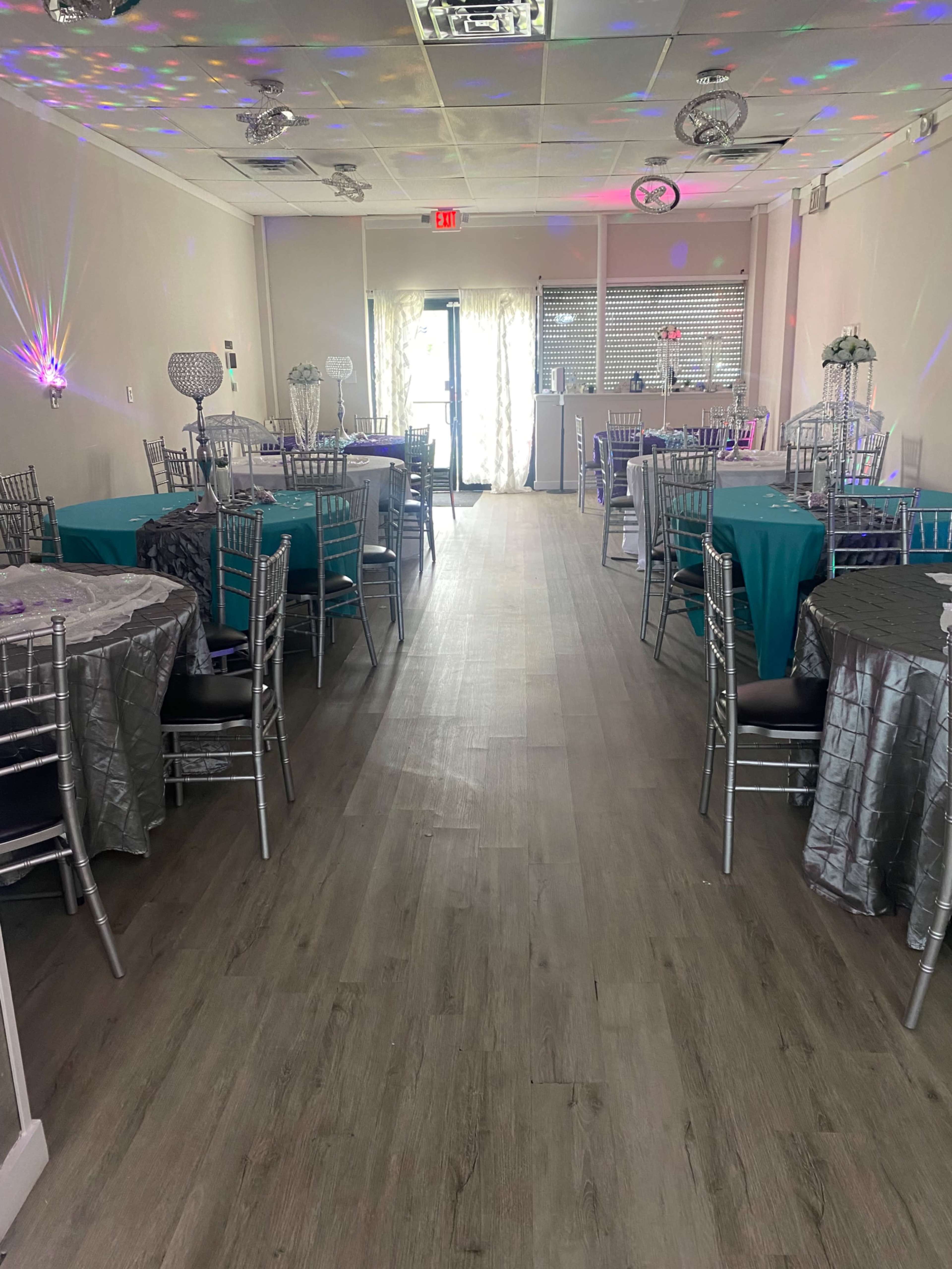 An event space with multiple tables dressed in teal and silver linens arranged neatly, leading to a doorway at the far end.