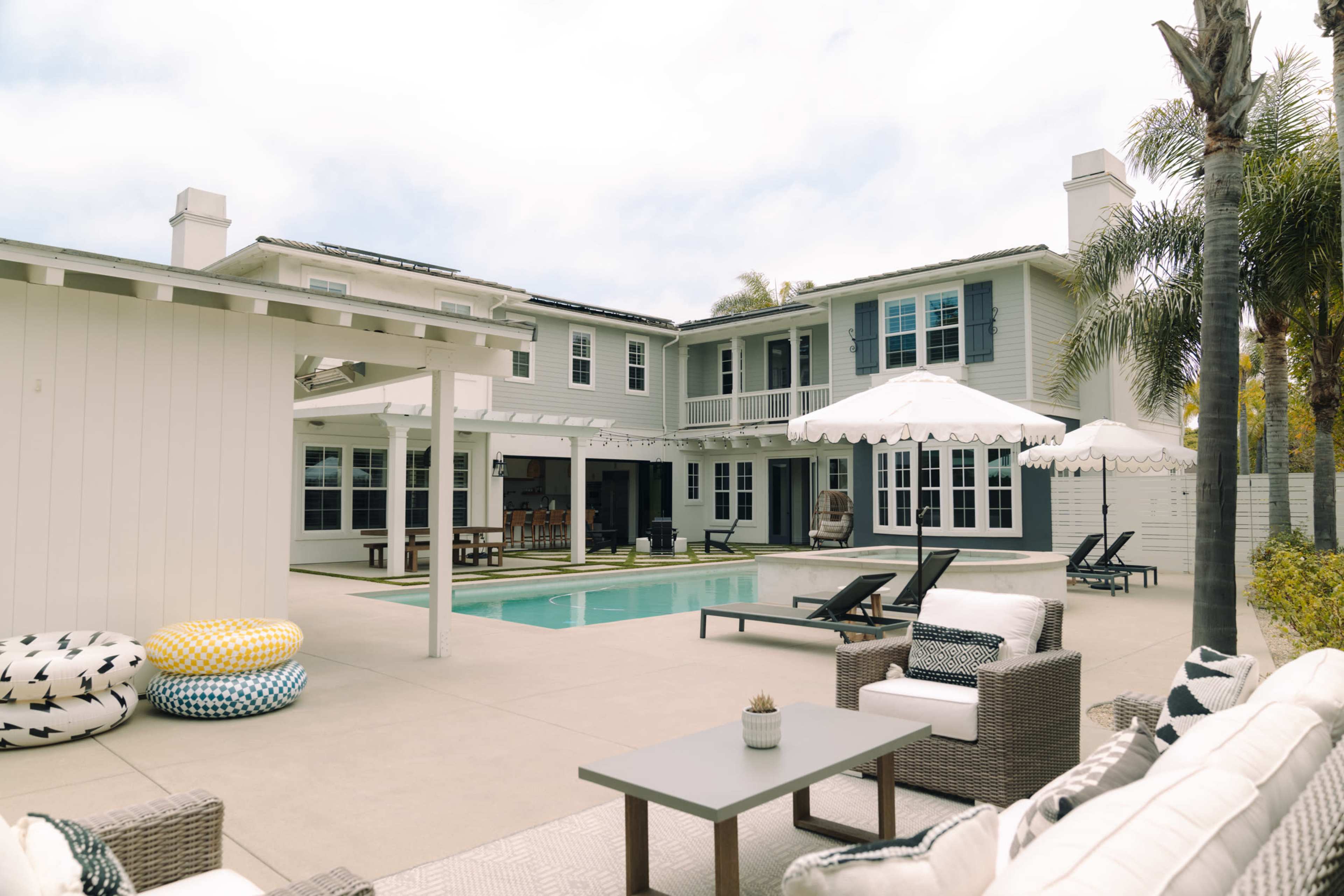 The image shows a modern backyard with a swimming pool, lounge chairs, and a seating area, surrounded by a two-story house and palm trees.