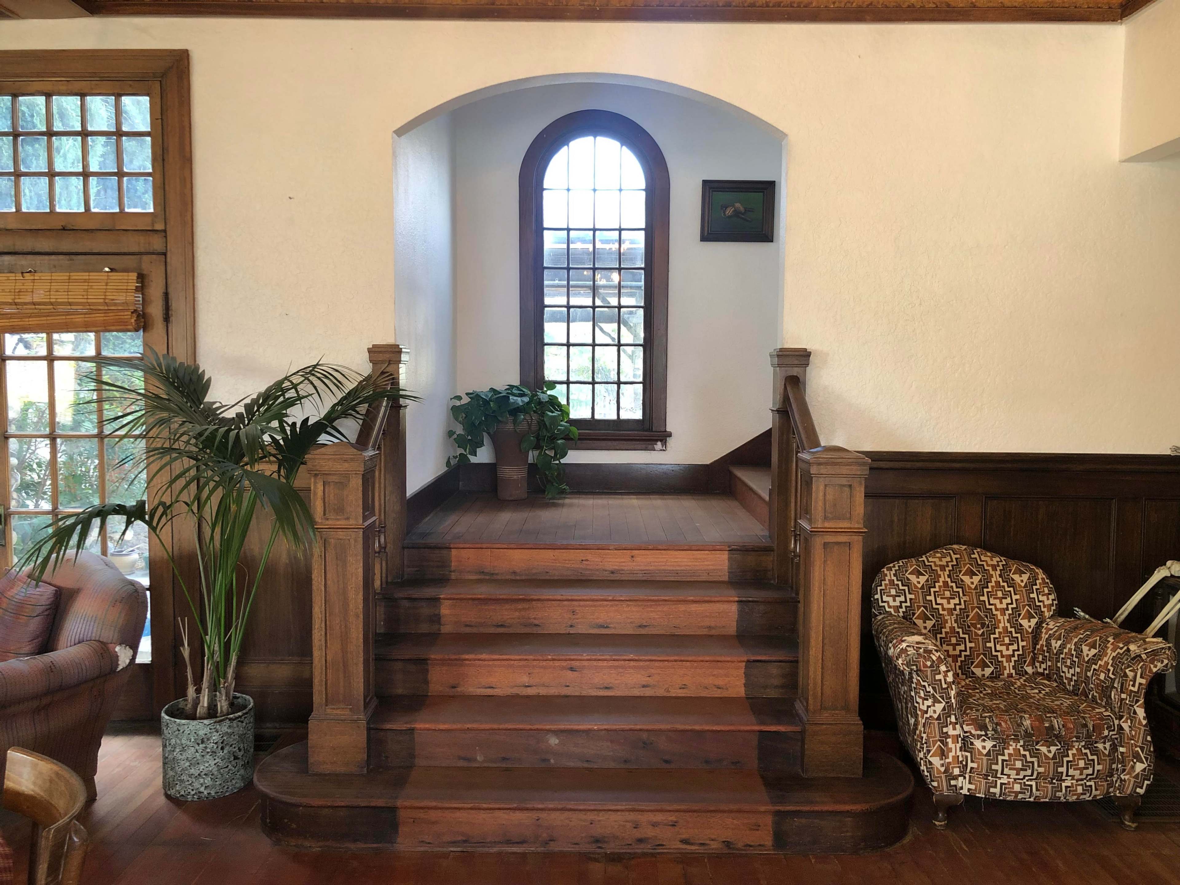 The image shows a staircase leading up to a landing with a large arched window and a potted plant on a ledge.