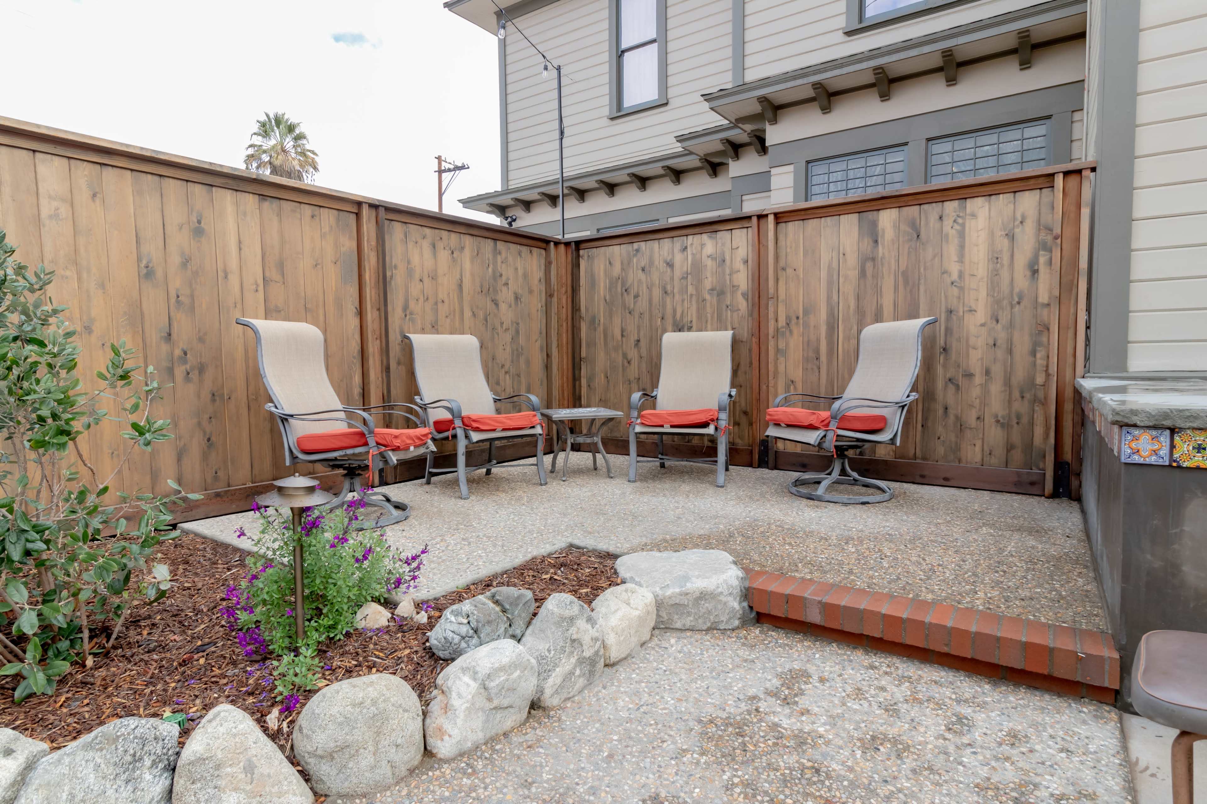 The image shows a small outdoor patio area with four lounge chairs and a circular table, surrounded by wooden fencing and landscaping features.