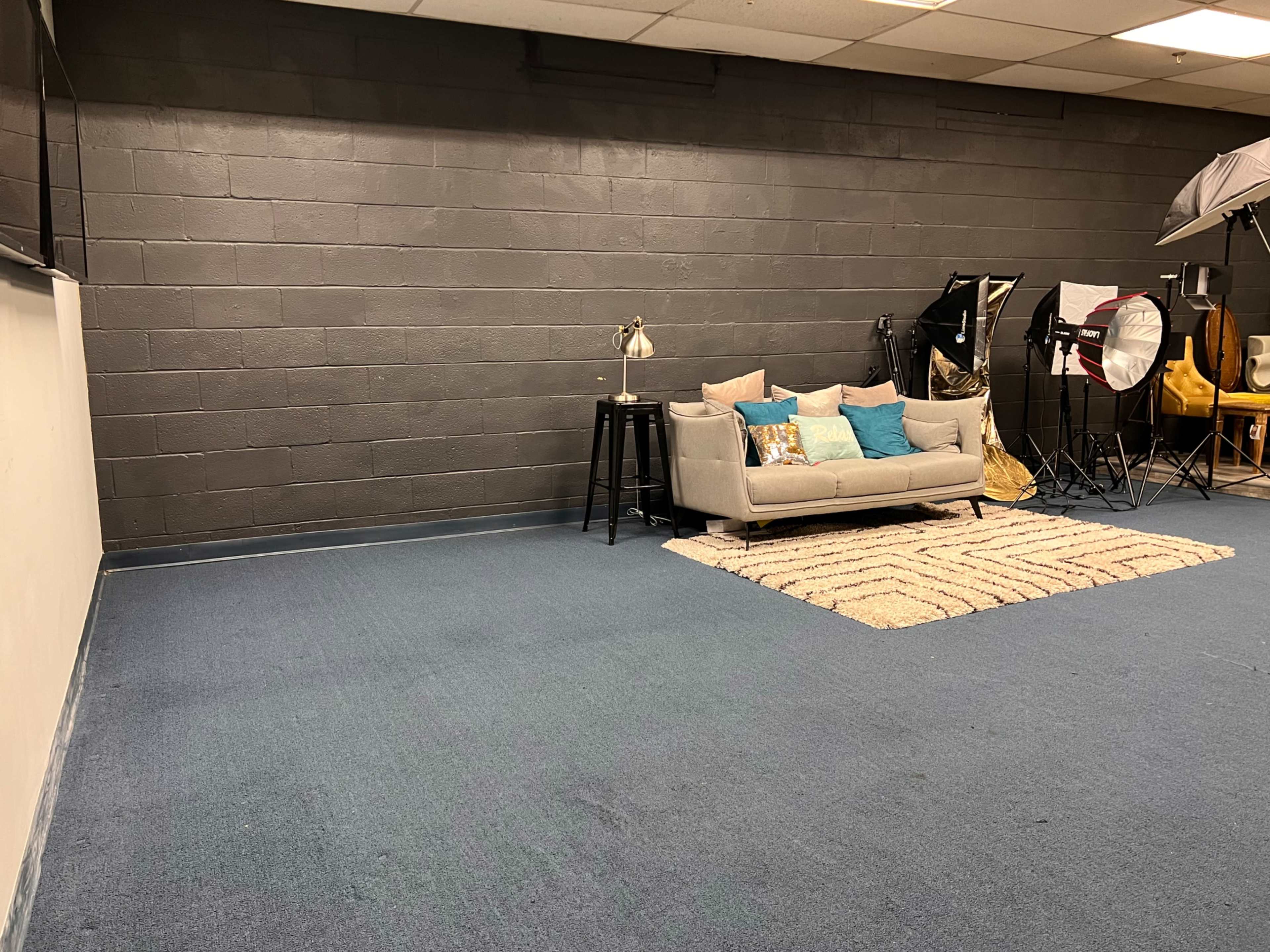 A spacious room with a gray wall, a light gray couch adorned with blue pillows, a small black side table with a lamp, and photography equipment on the right side.