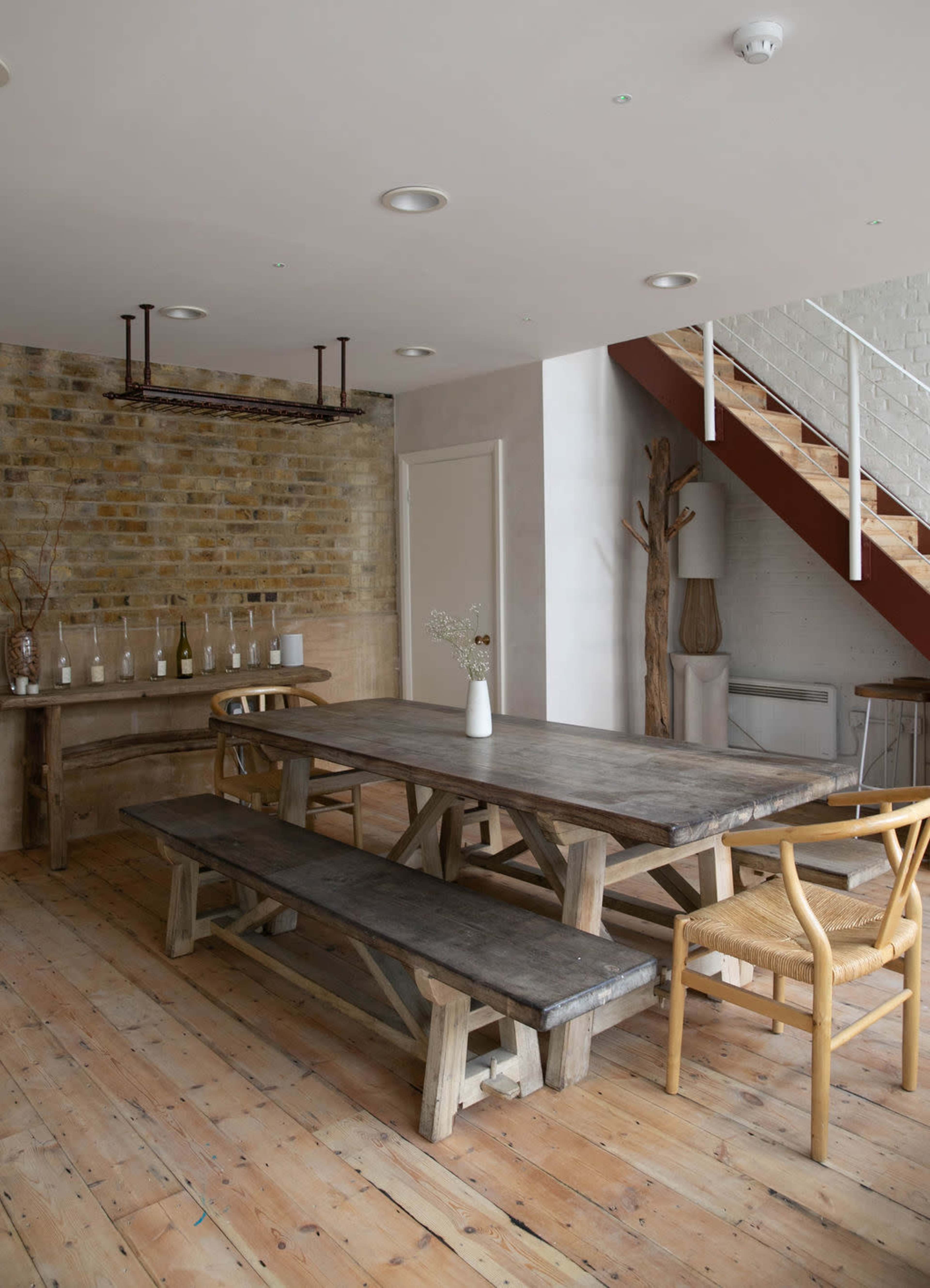 A spacious dining area features a large wooden table with matching benches, set against a brick wall, and a staircase in the background.
