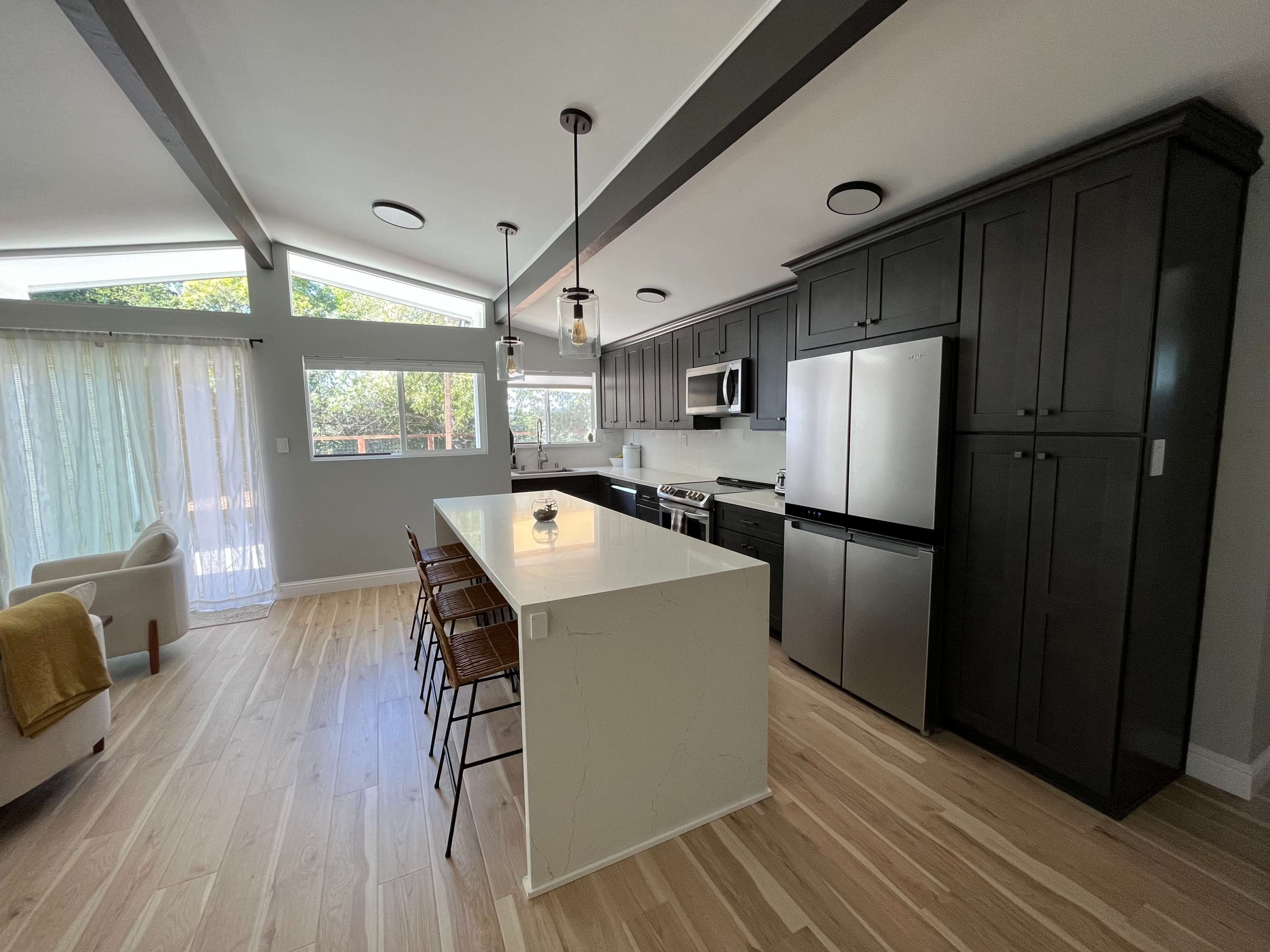 A modern kitchen with dark cabinets, a large island with bar stools, stainless steel appliances, and windows that allow natural light to enter the space.