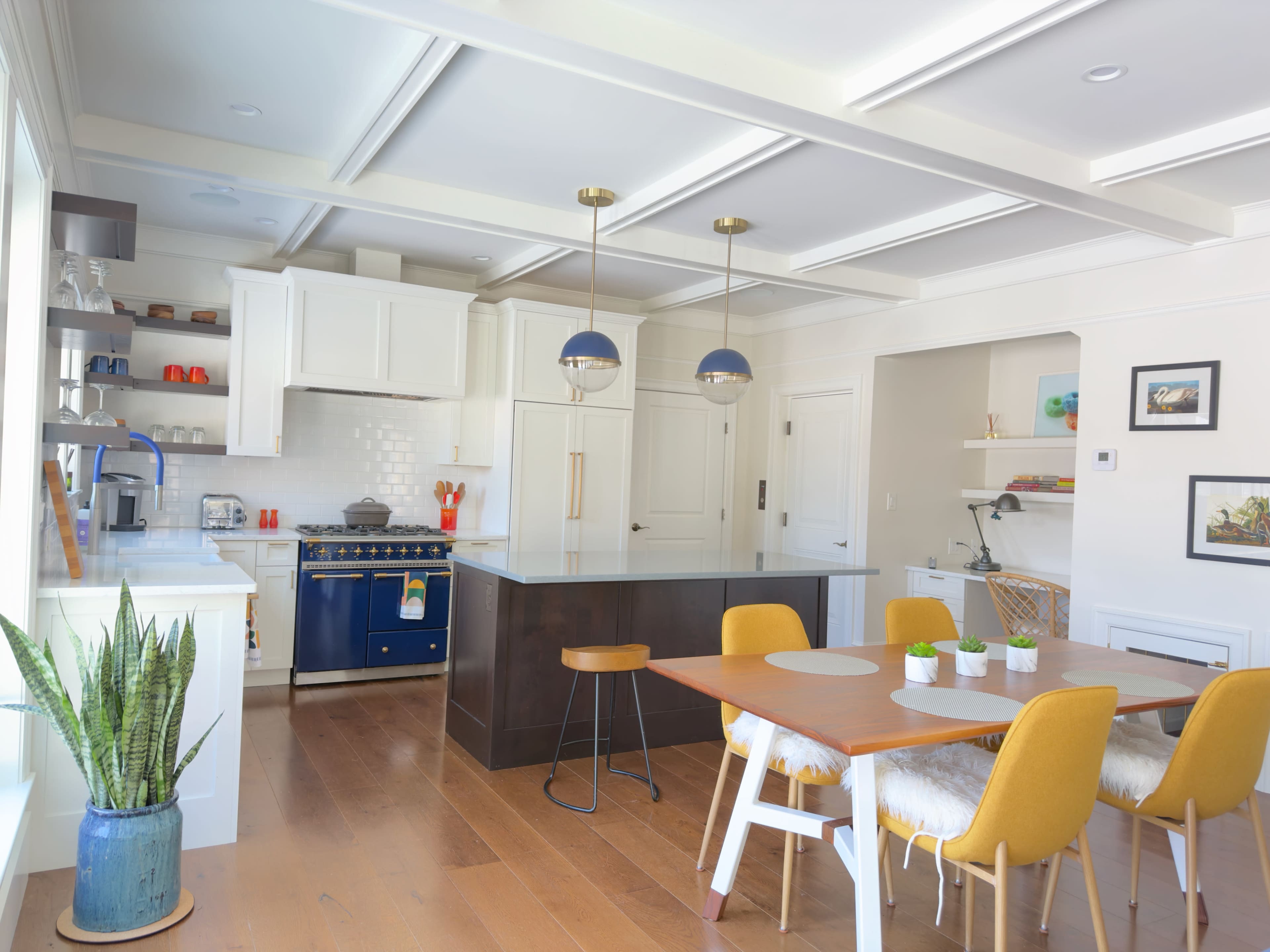 The image shows a modern kitchen with a large island, featuring blue cabinetry, a dining area with a wooden table and yellow chairs, and various plants and decorative items.