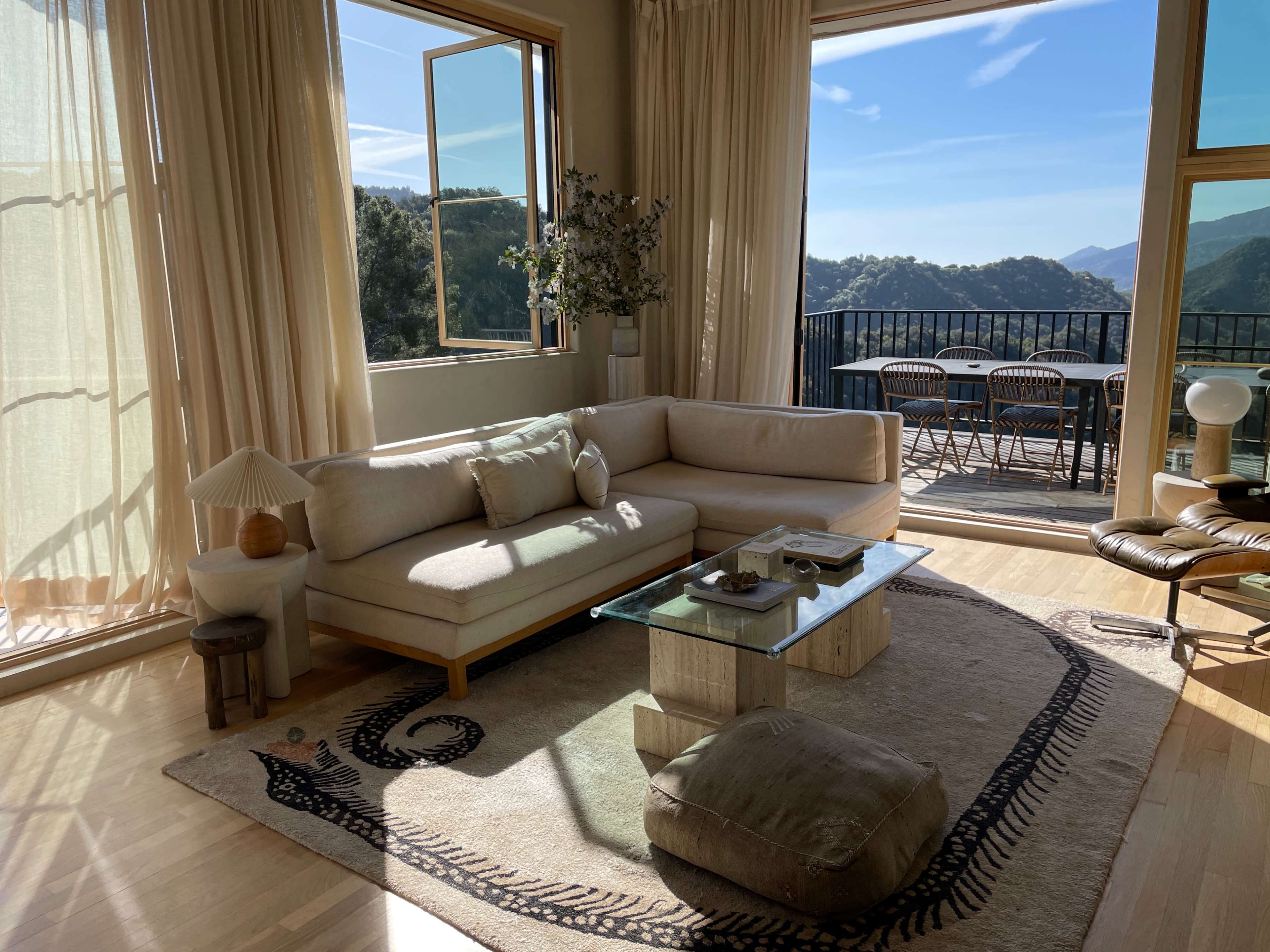 The image features a modern living room with large windows overlooking a mountainous landscape, furnished with a light-colored sofa, a glass coffee table, and a textured area rug.