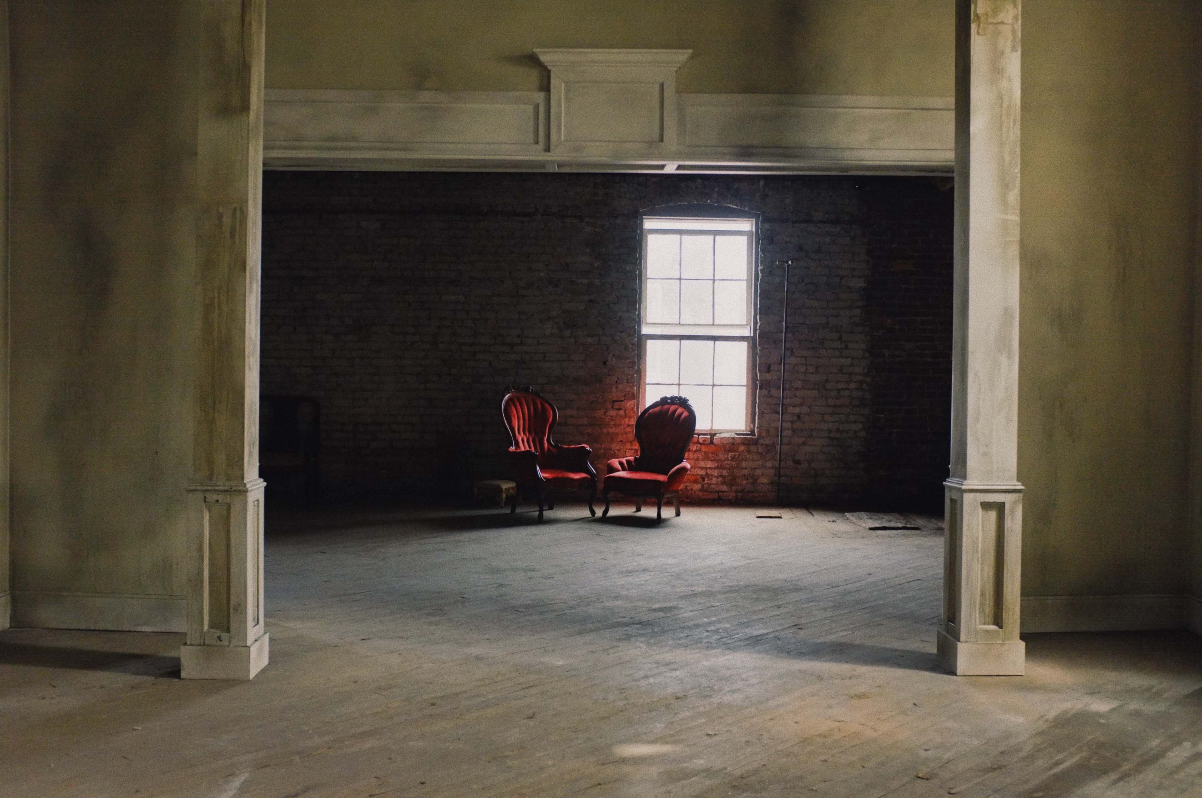 A pair of red chairs is positioned in a dimly lit room with exposed brick walls and a large window.