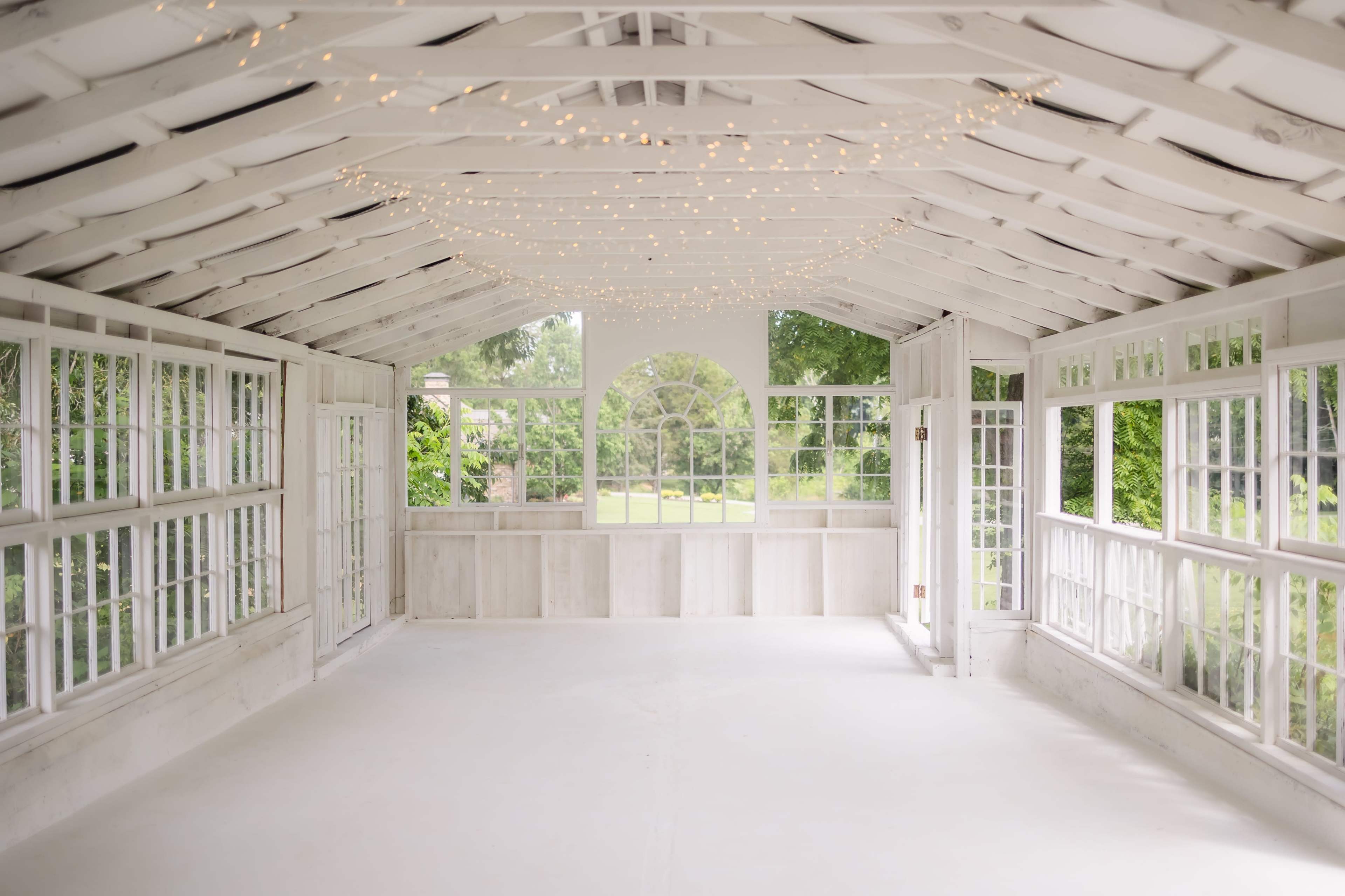 The image shows a bright, empty indoor space with white walls and ceiling, large windows, and string lights along the roof, overlooking a garden.