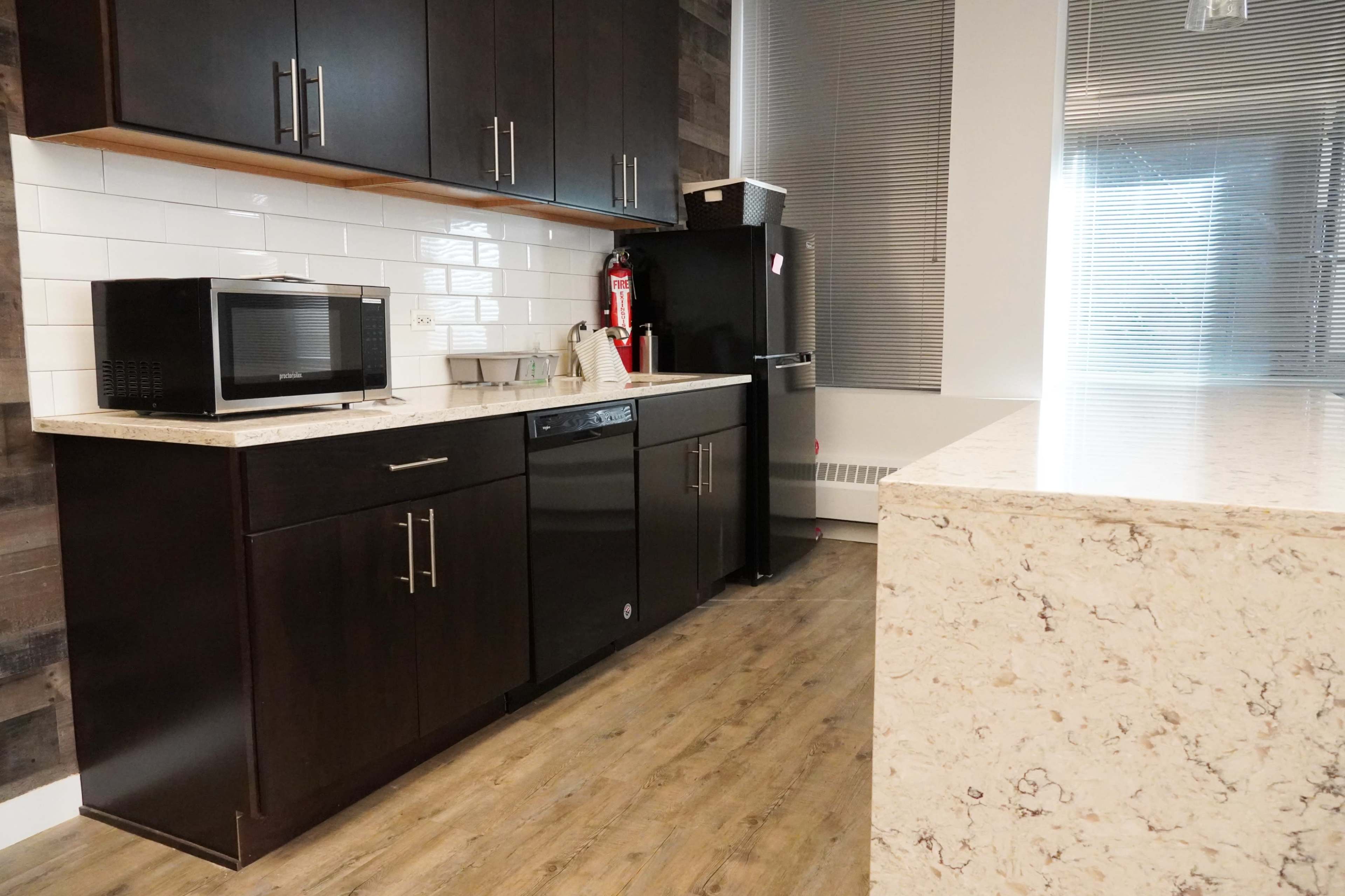 A modern kitchen features dark cabinetry, a countertop, a microwave, and a dishwasher, with a refrigerator in the corner and large windows providing natural light.
