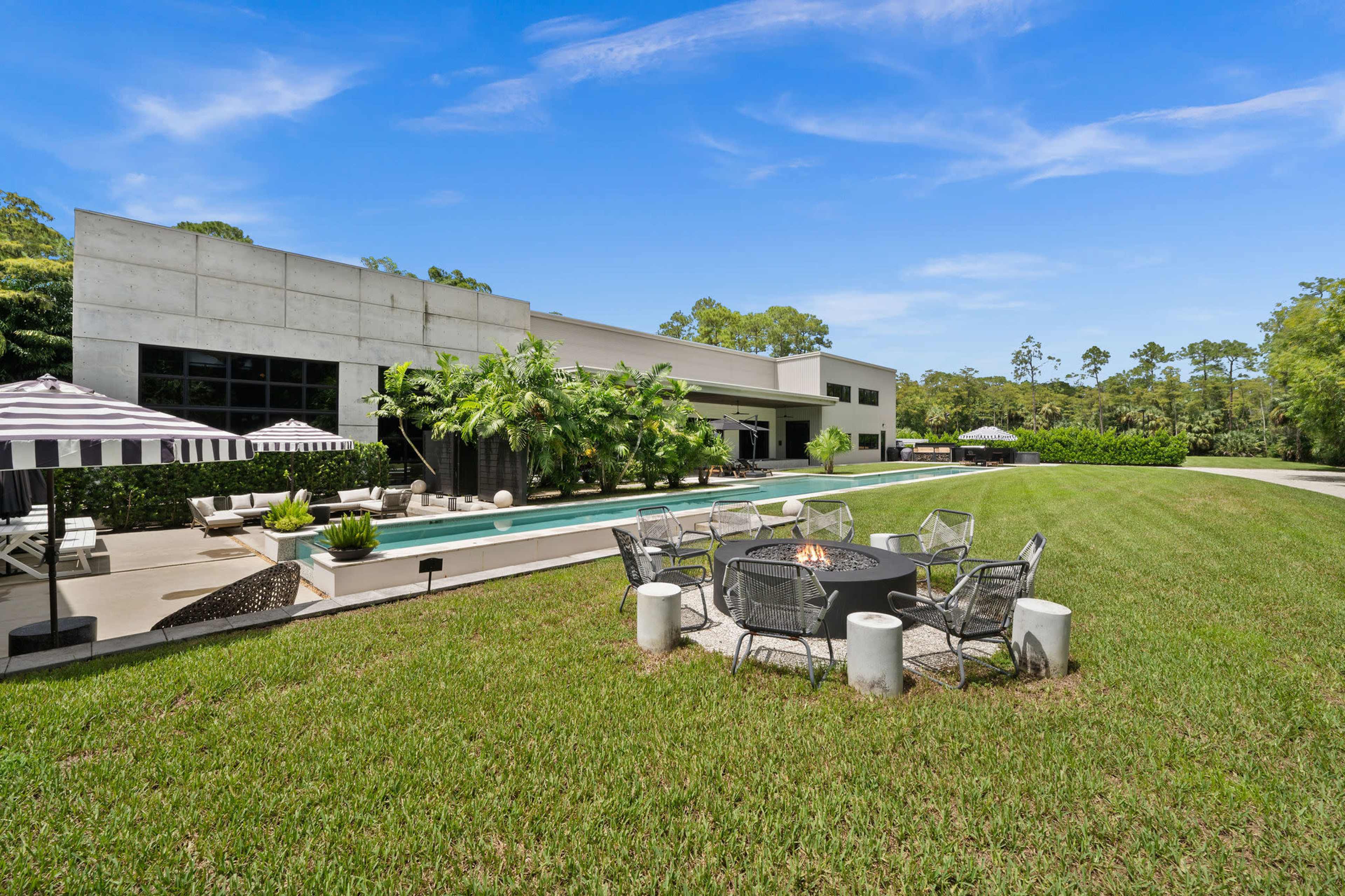 A modern house with a large swimming pool is surrounded by green grass and features an outdoor seating area with a fire pit.