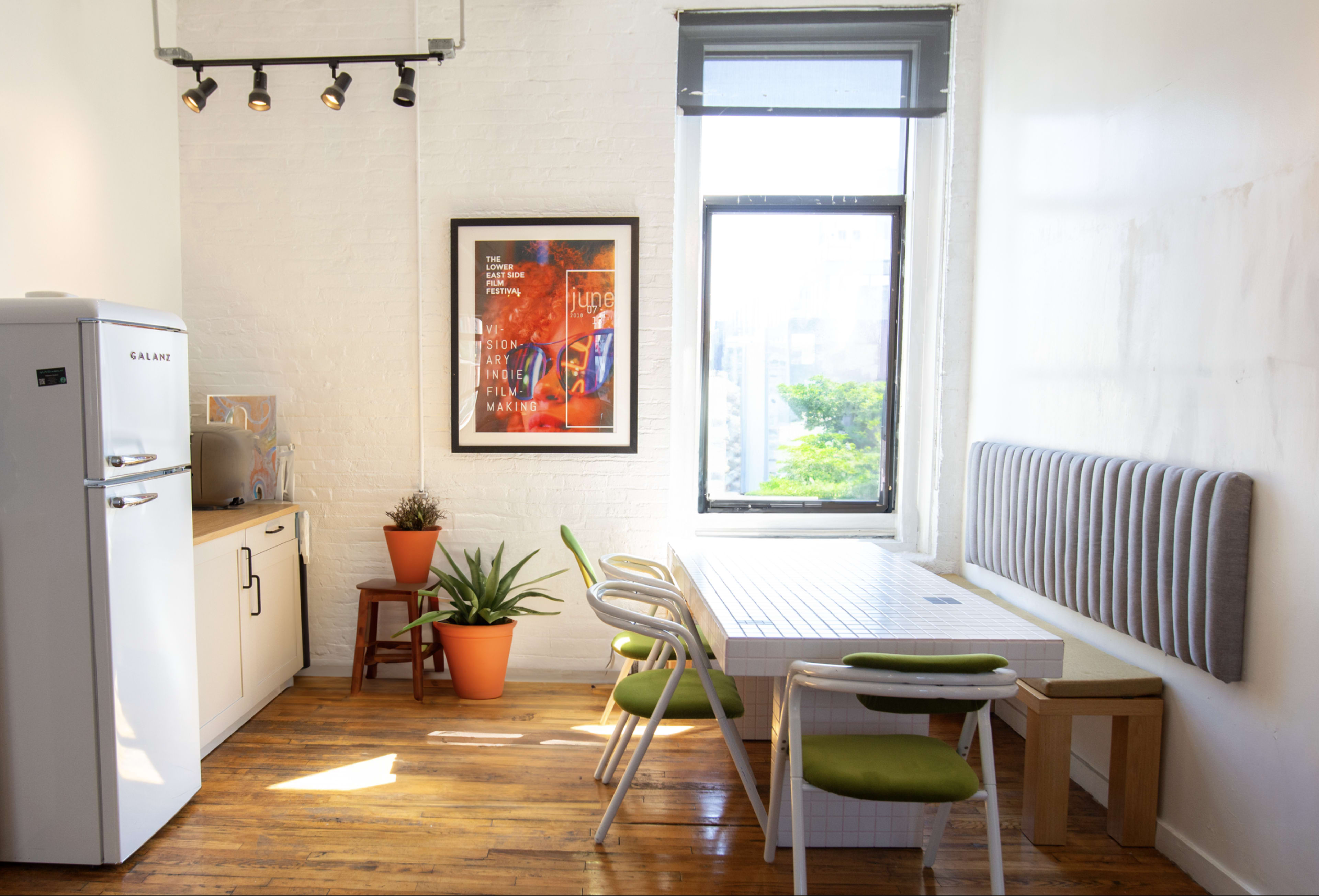A modern kitchen area features a white refrigerator, a dining table with green chairs, and a large window with a view of greenery outside.