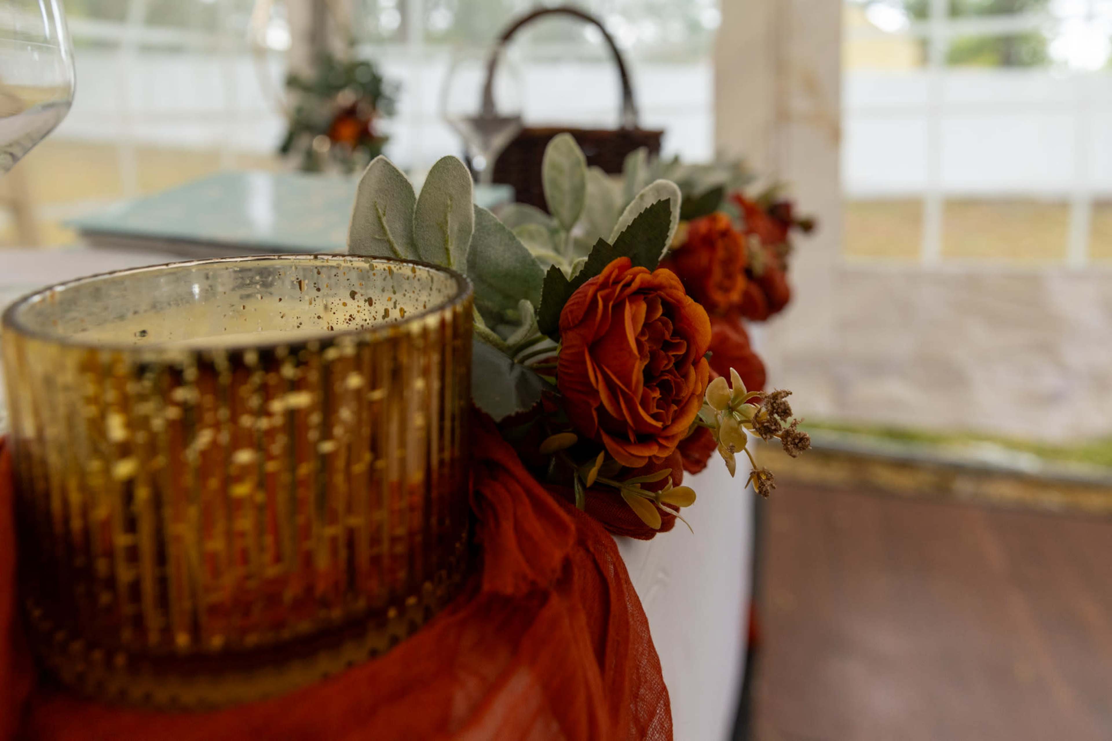 A decorative arrangement features a gold candle holder and artificial flowers in shades of orange and green, set against a backdrop of a tented event space.